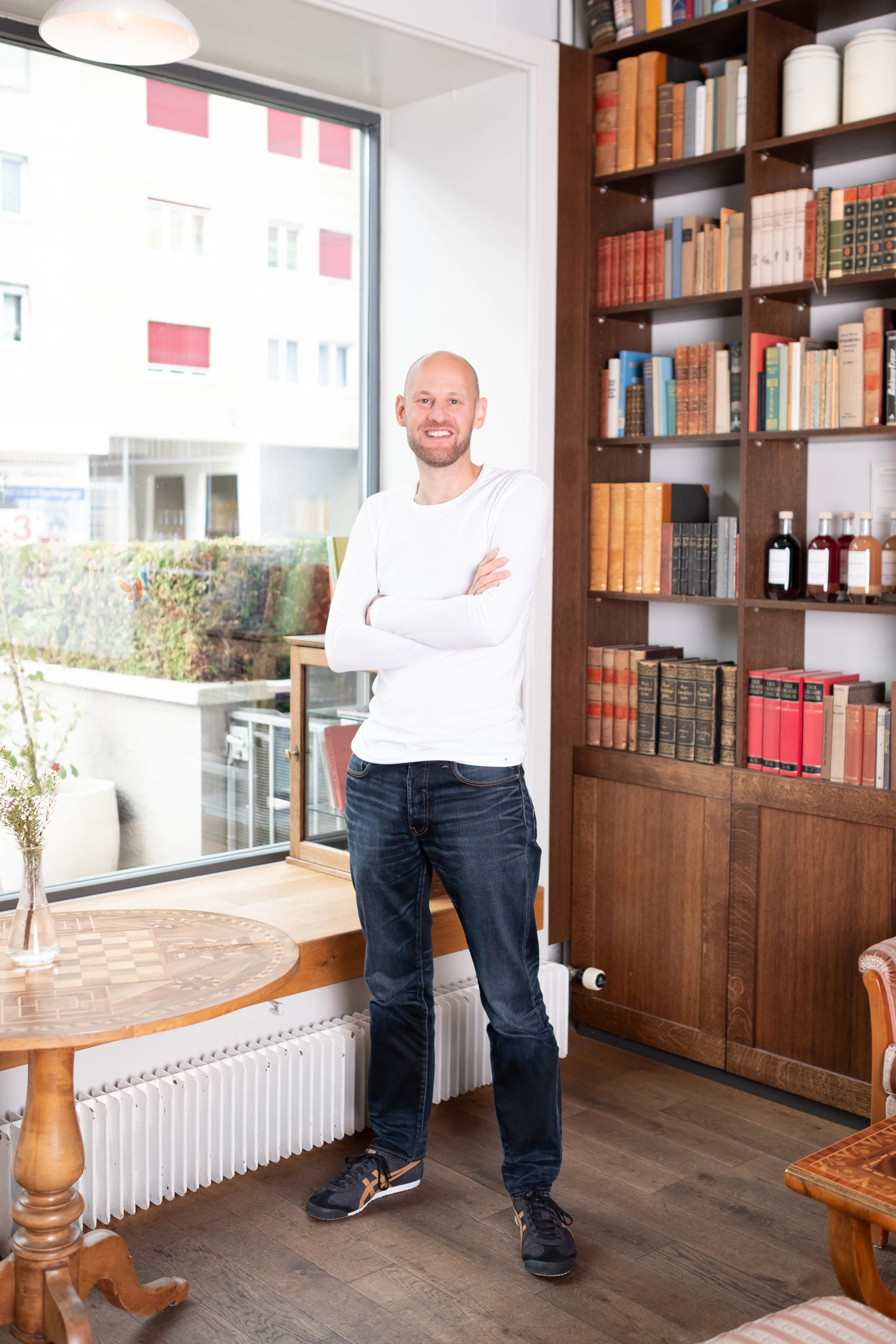 Ein lächelnder Mann steht mit verschränkten Armen in einem Raum mit Bücherregalen aus Holz, großen Fenstern und Holzboden.