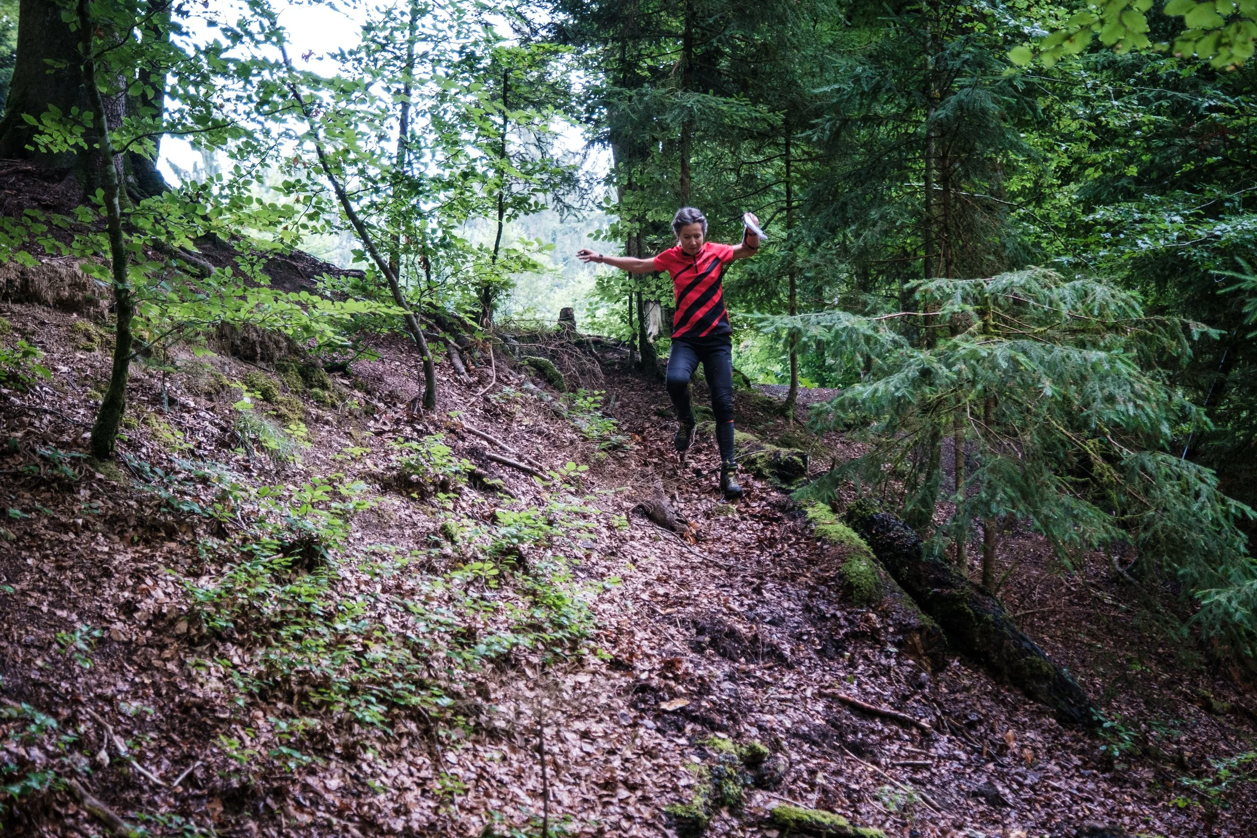 Eine Frau in einem Wald, die einen steilen, schlammbeladenen Pfad hinunterrutscht oder -springt.