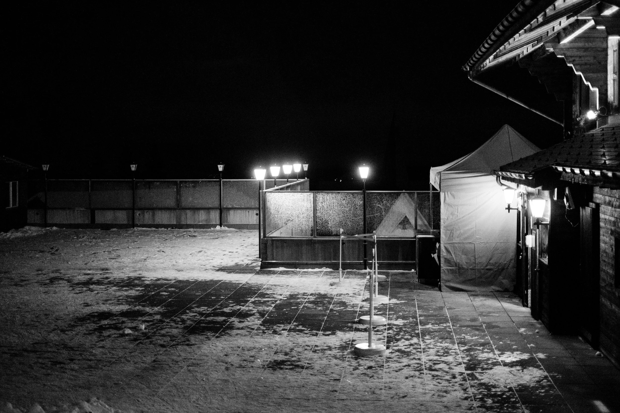 Schneebedeckter Außenbereich bei Nacht mit Zaun, Beleuchtung und einem Zelt, dunkler Himmel.