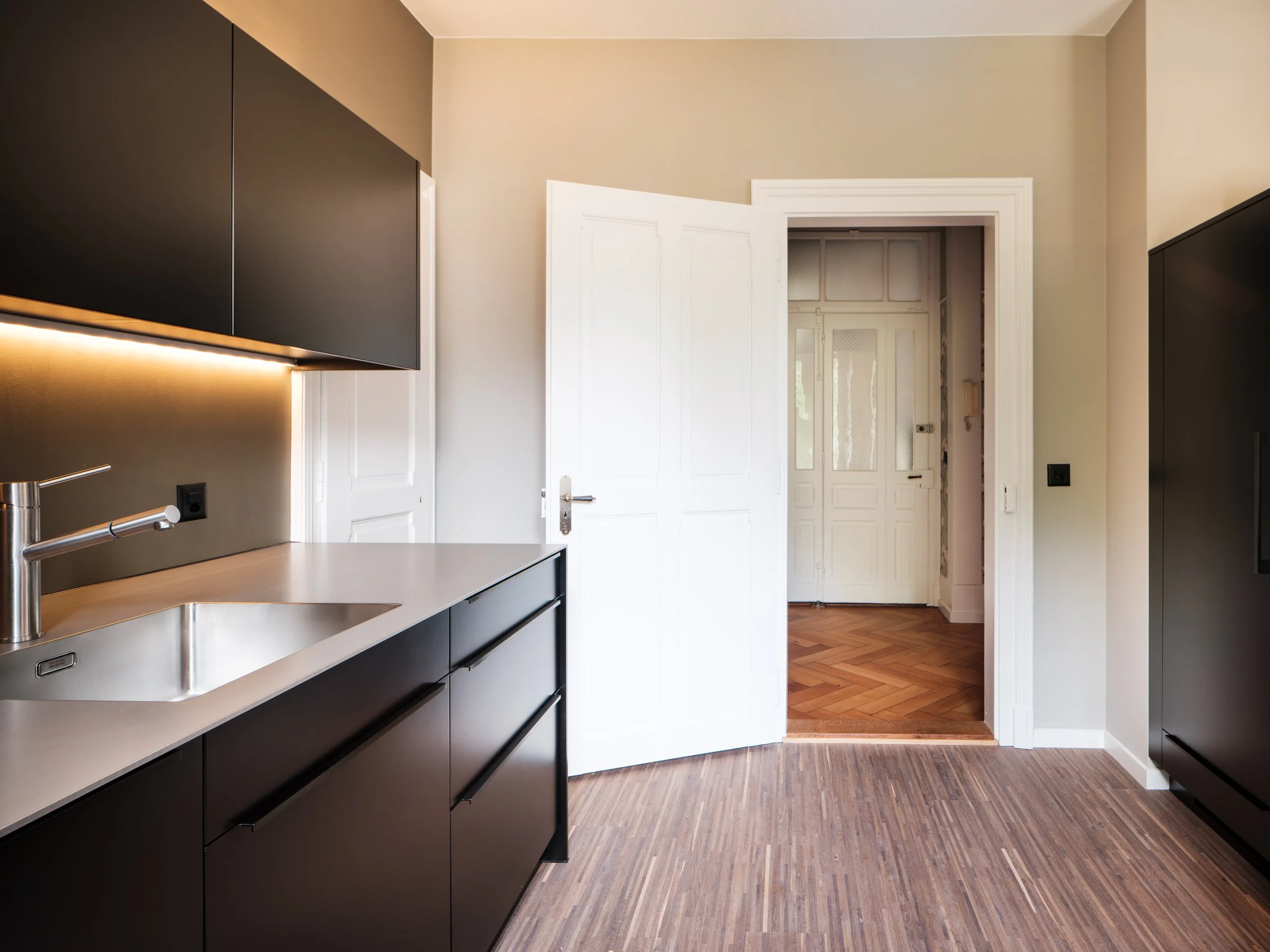 Moderne schwarze Küchenschränke mit weißen Arbeitsflächen, Edelstahlspüle, offenes weißes Tür und Dielenboden.