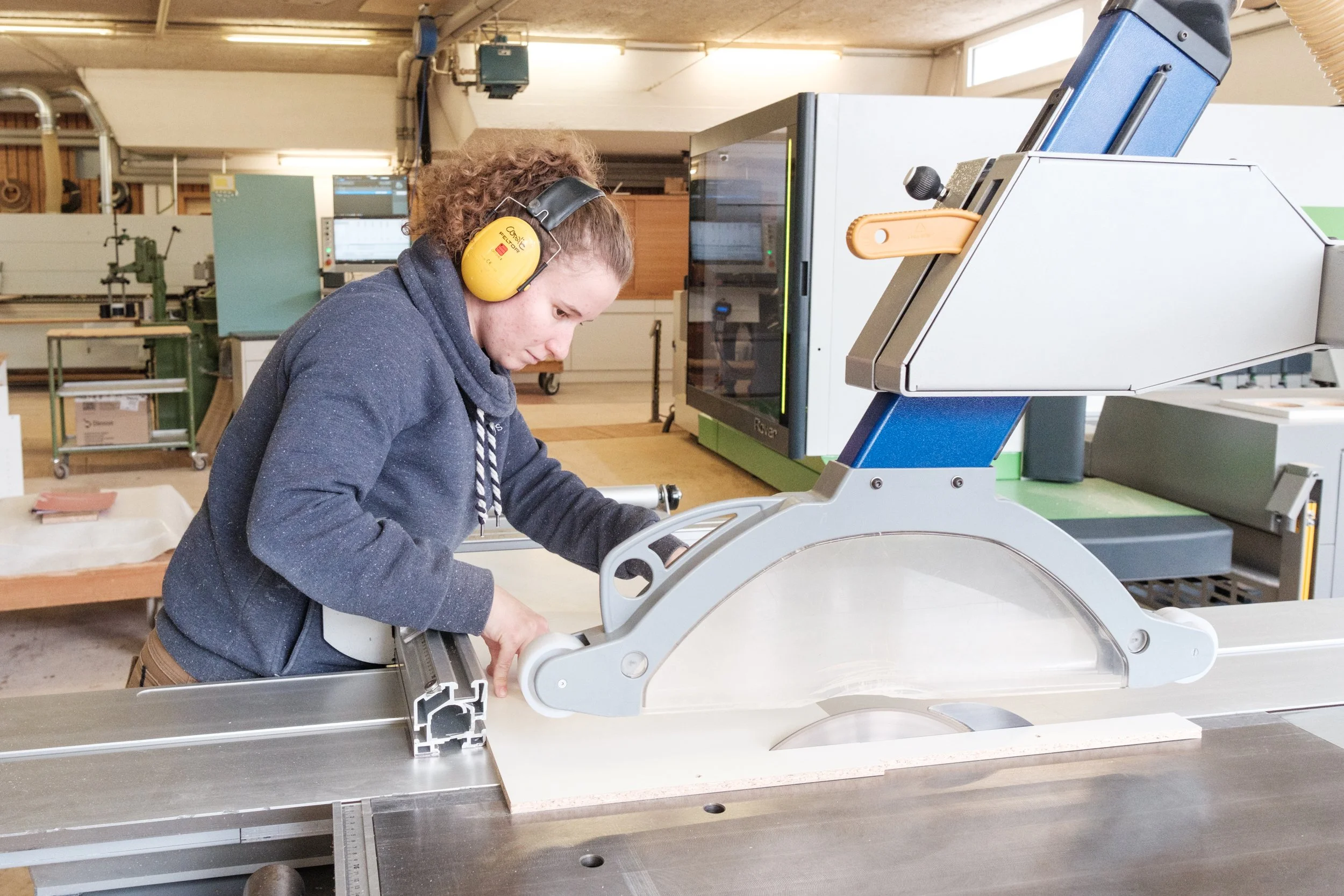 Mensch arbeitet in einer Werkstatt mit einer Tischkreissäge, trägt Gehörschutz und konzentriert sich auf die Arbeit.