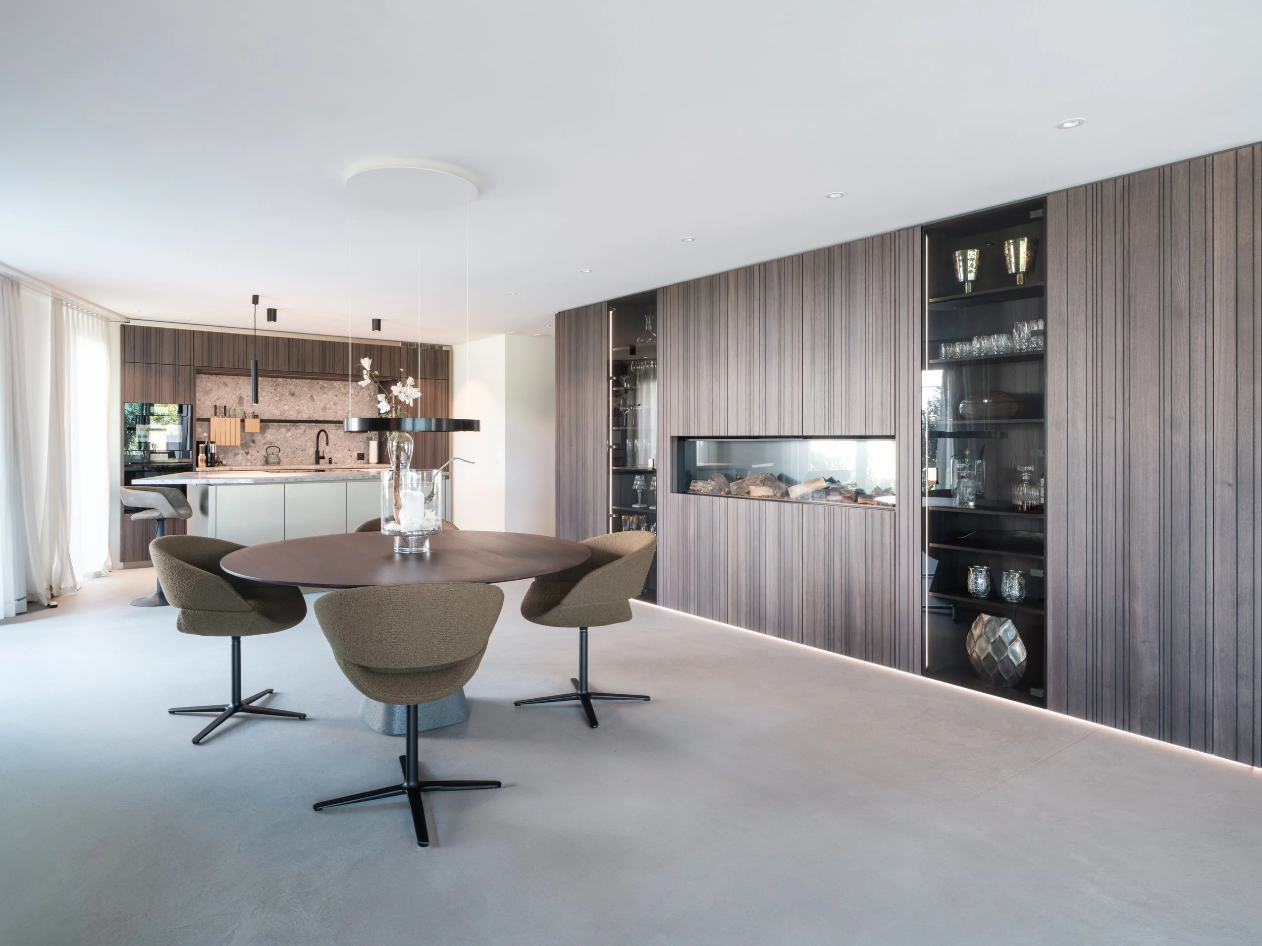 Moderner Speisesaal mit runder Holz-Tisch, sechs bequemen, braunen Sesseln, dunkler Holzverkleidung an den Wänden und eingebautem Kaminsystem, große Fenster mit weißen Vorhängen, minimalistischer Kronleuchter und eine offene Küche im Hintergrund.