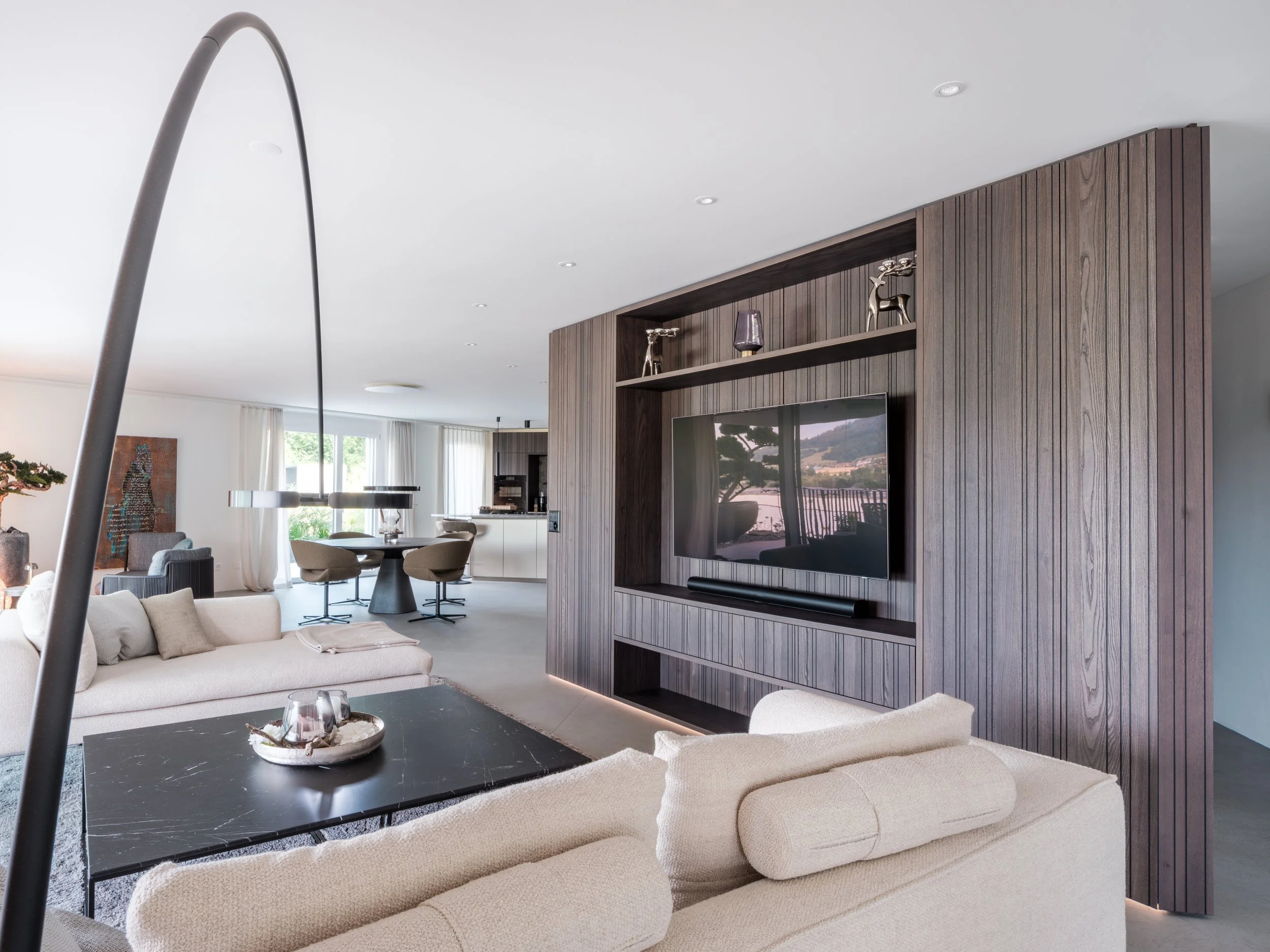 ModernerWohnraum mit beige Couch, schwarzem Couchtisch, TV in Holz-Wandverkleidung, offenen Küche, Essbereich mit runden Tisch und Stühlen, große Fenster mit Vorhängen.