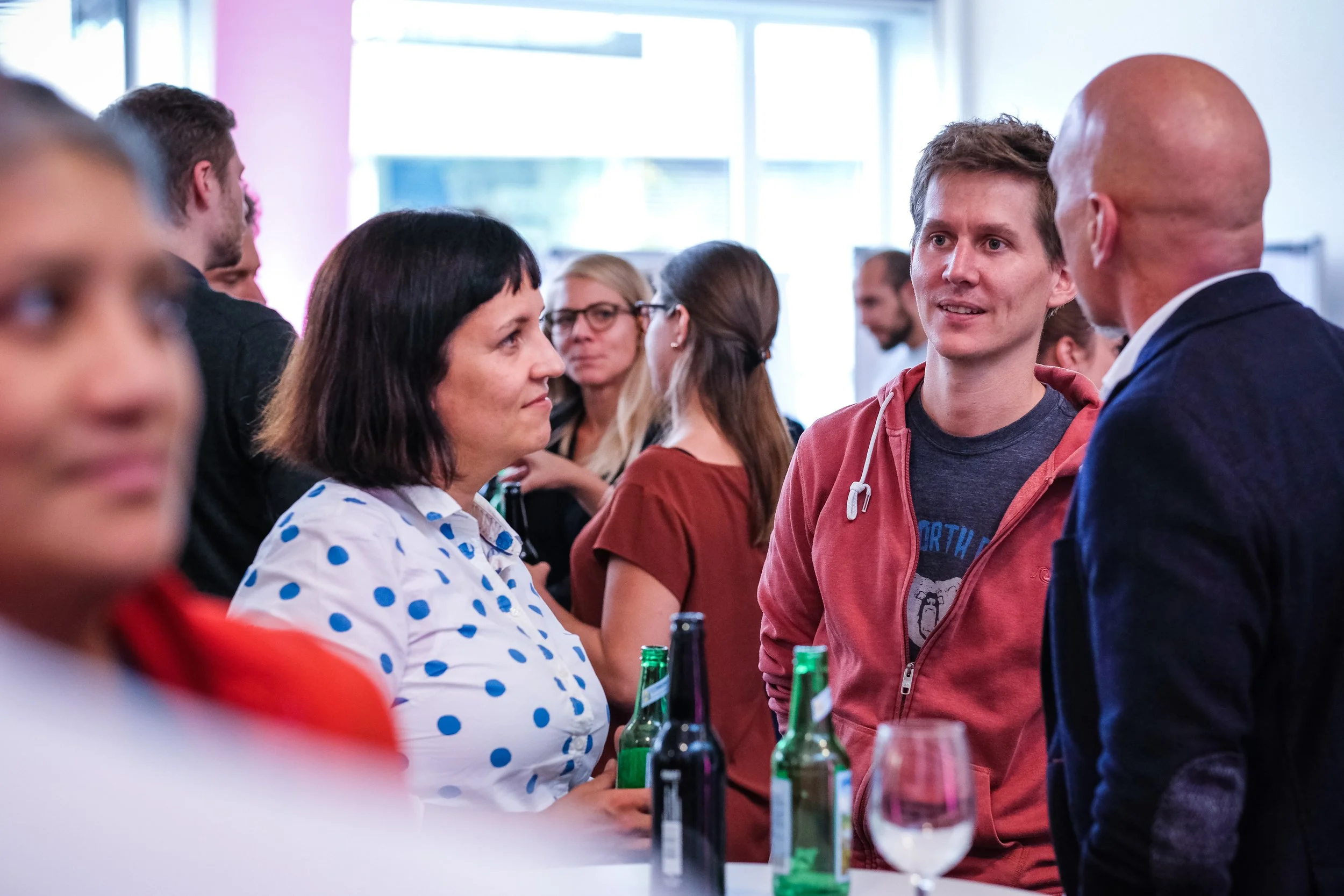 Menschen beim Gespräch auf einer Party oder in einem sozialen Umfeld, Getränke auf dem Tisch, im Hintergrund weitere Personen, helle Fensterfront