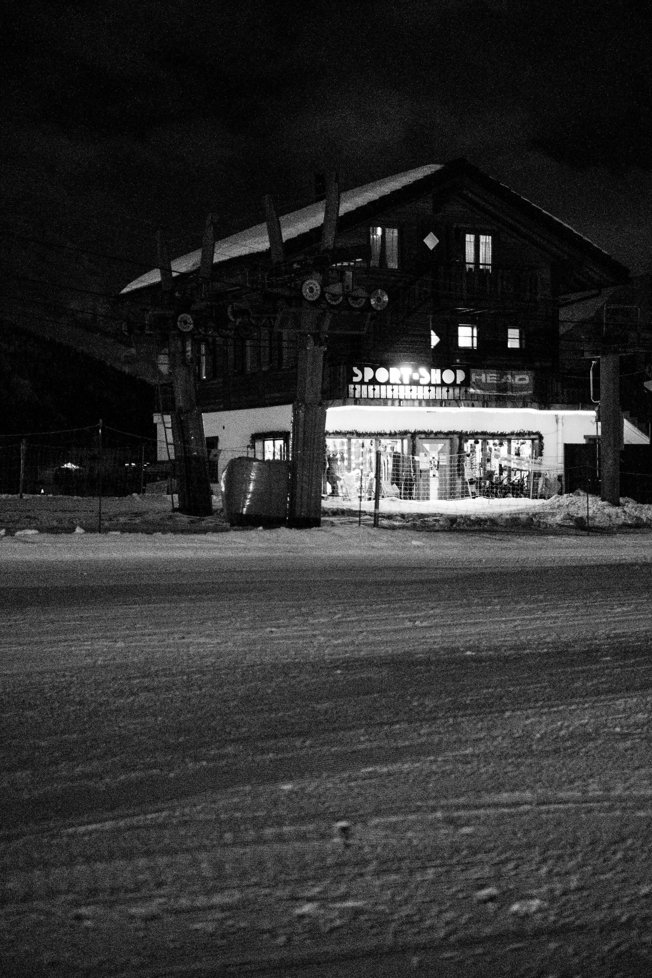 Schneebedecktes Gebäude bei Nacht, mit einem Laden namens "Sport-Shop" im Erdgeschoss, flackerndes Licht, dunkler Himmel, verschneiter Straßenbelag im Vordergrund.