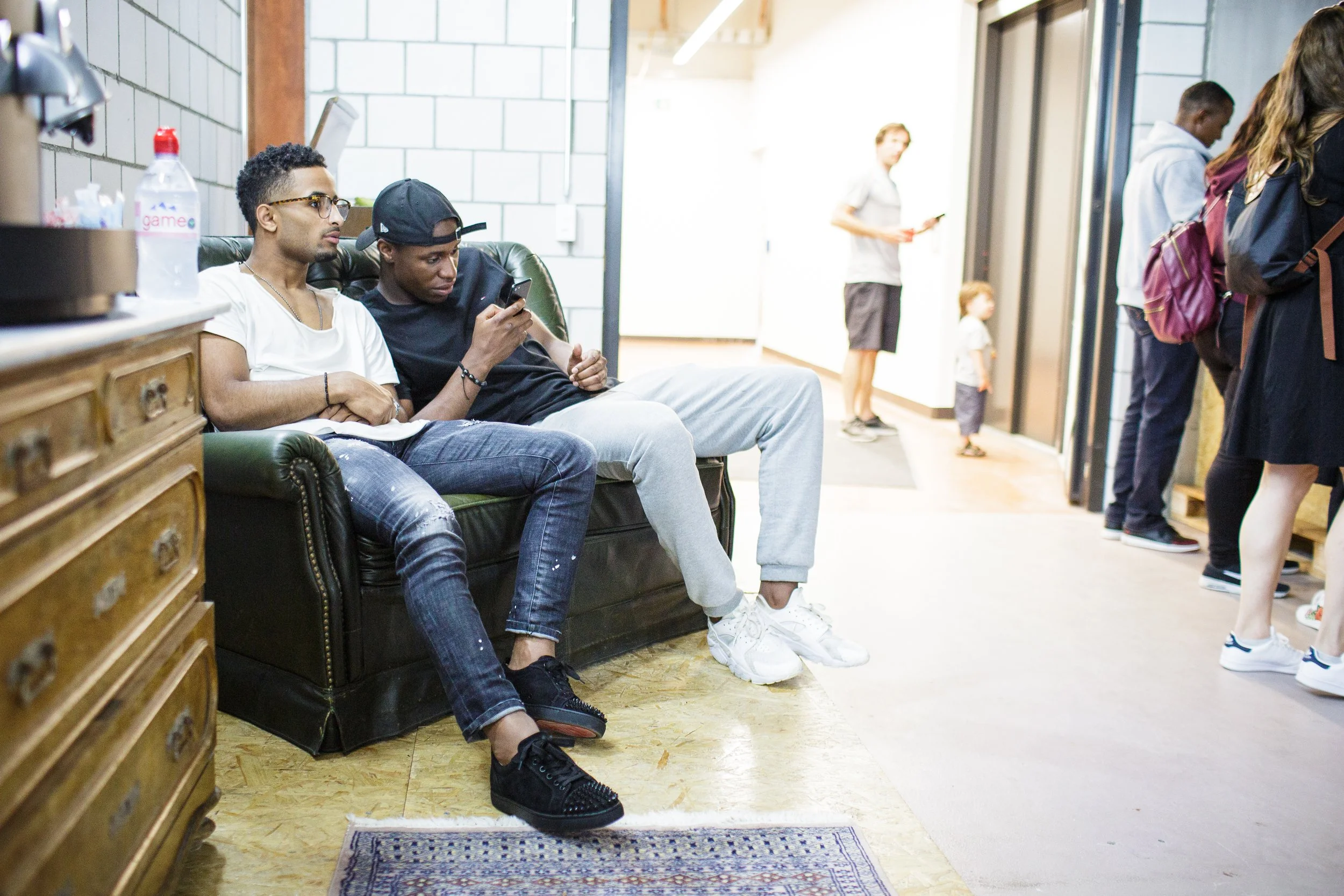 Zwei junge Männer sitzen auf einer grünen Couch, einer liest und der andere schaut auf sein Smartphone, im Hintergrund ziehen weitere Menschen durch einen Flur.