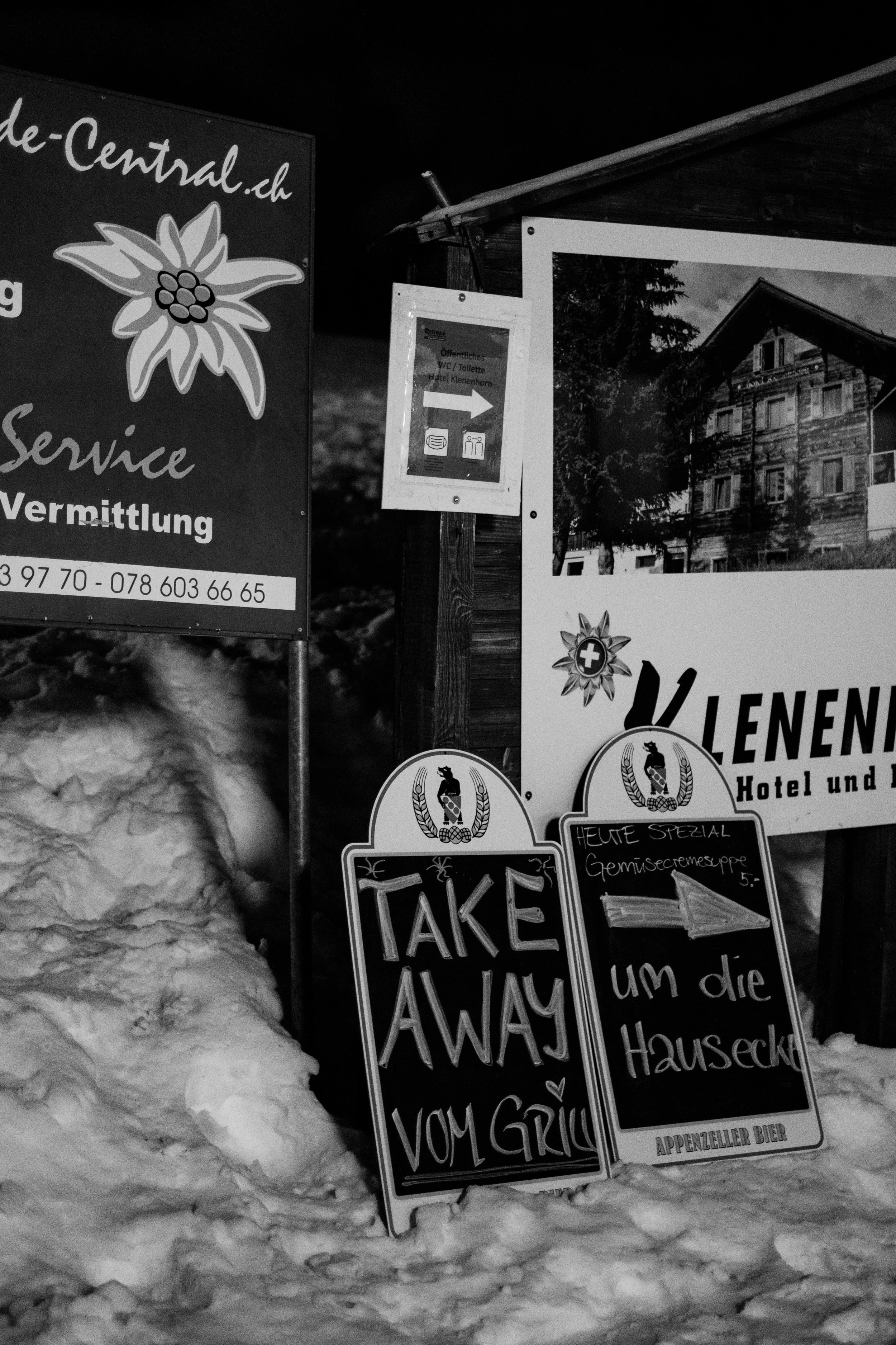 Schilder vor einem Hotel in Schneelage, eines zeigt 'Take Away vom Grill', ein anderes bewirbt eine Gemüsesuppe und weitere Hinweise, alles in Deutsch.