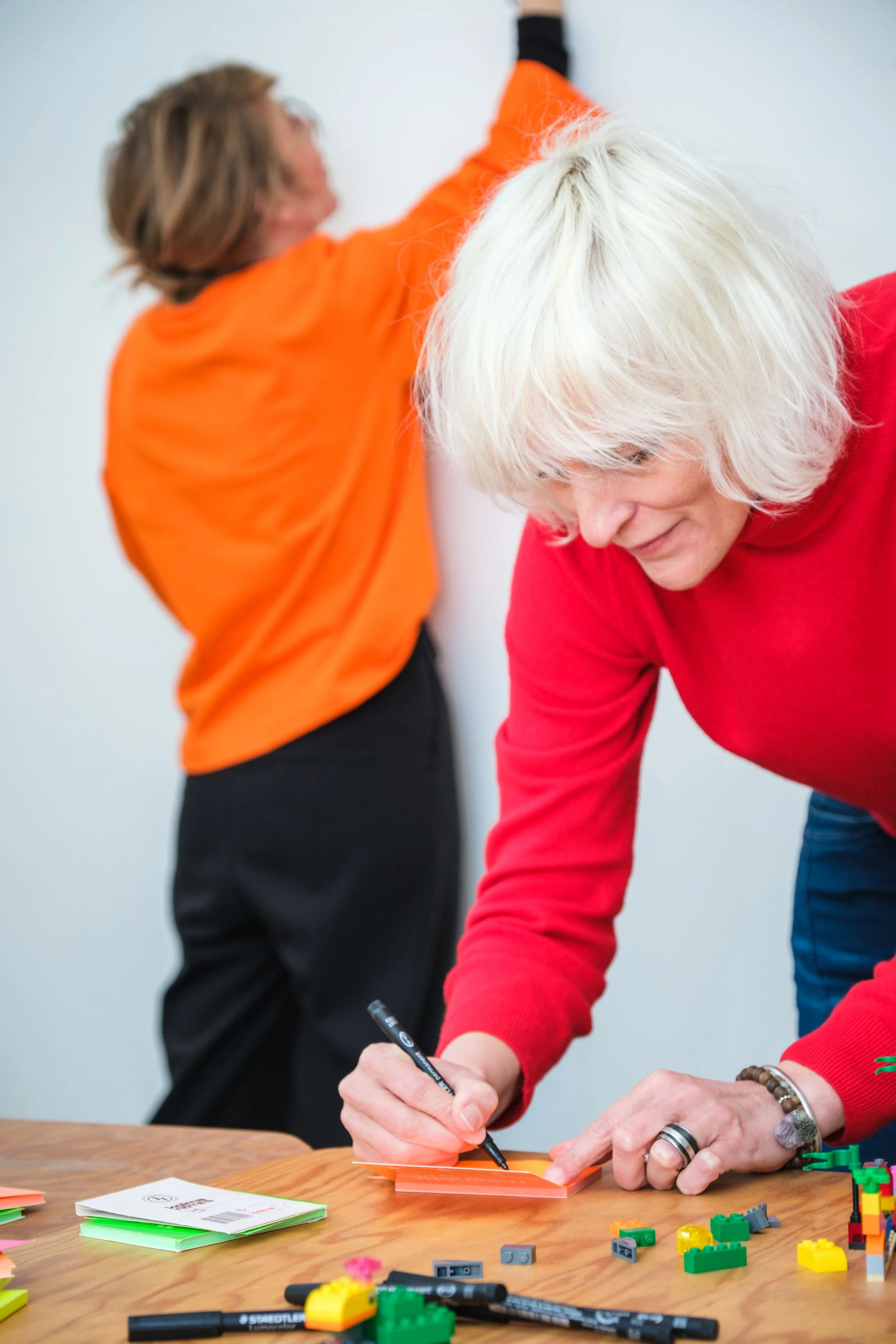 Zwei Frauen beim Schreiben und Spielen mit Legosteinen an einem Tisch, im Hintergrund unklar, eine Frau trägt ein rotes Oberteil, die andere ein orangefarbenes Oberteil.