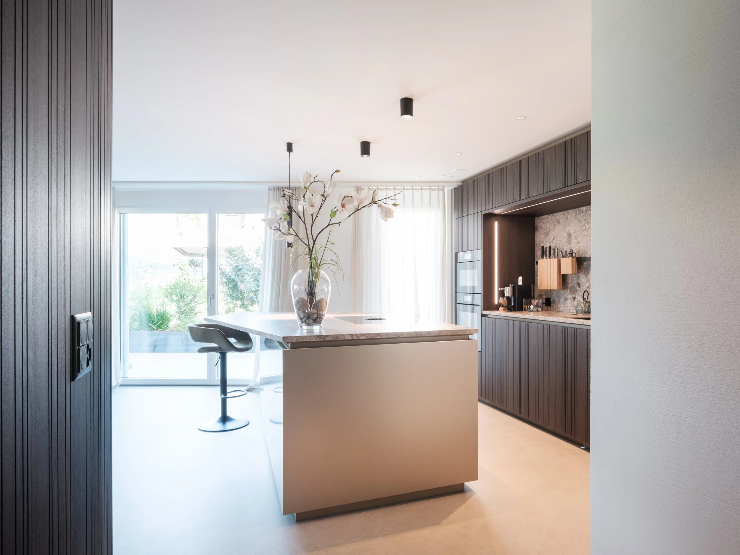 Moderner Küchenbereich mit dunklen Holzverkleidungen, einer Kücheninsel mit Blumenvasen, Kühlschrank, Kaffeezubehör und großen Fenstern mit weißen Vorhängen.