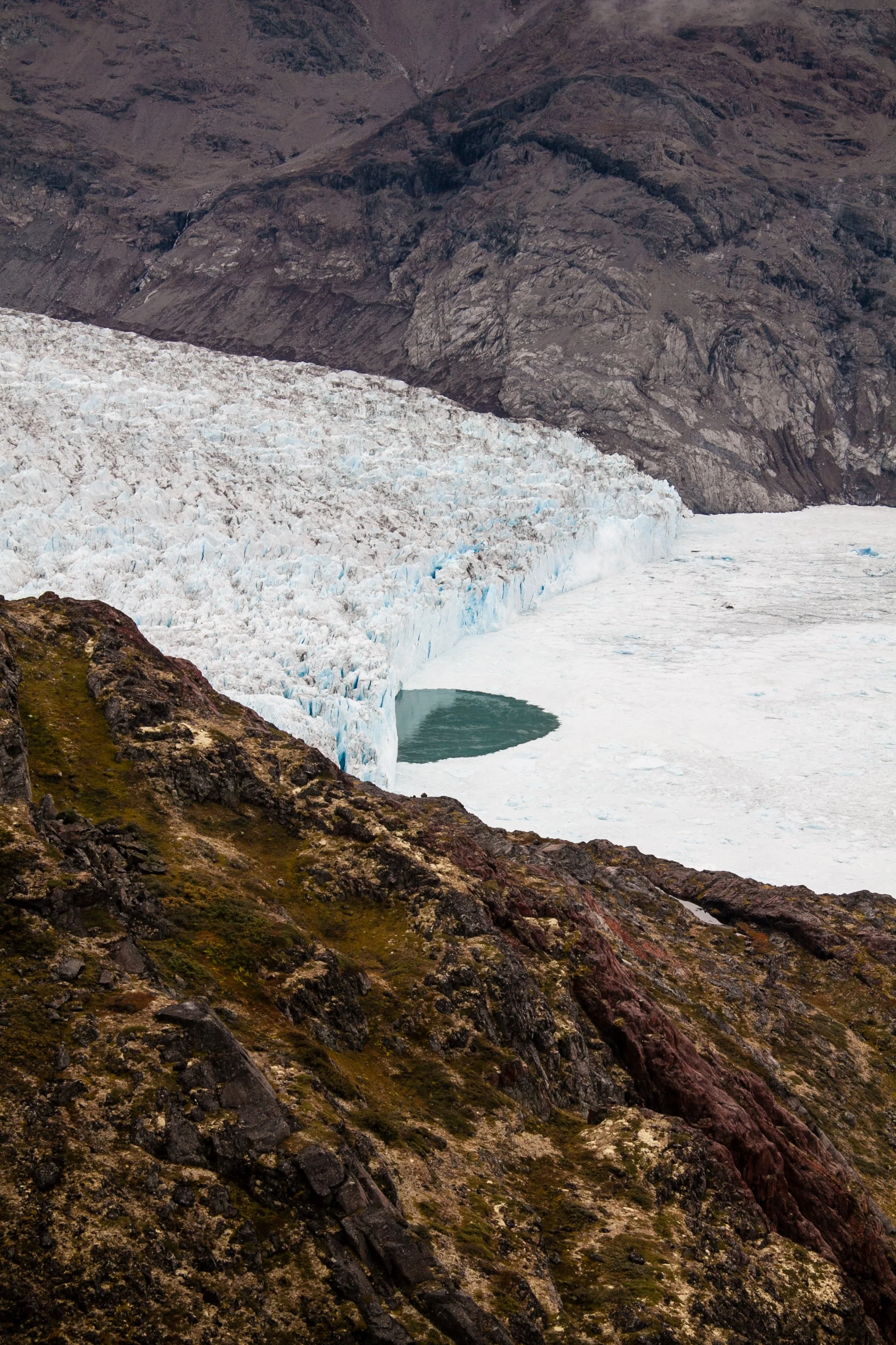 Gletscher mit Wasserloch in einem Fjord, umgeben von felsigen, grünen Hügeln und Bergen im Hintergrund.