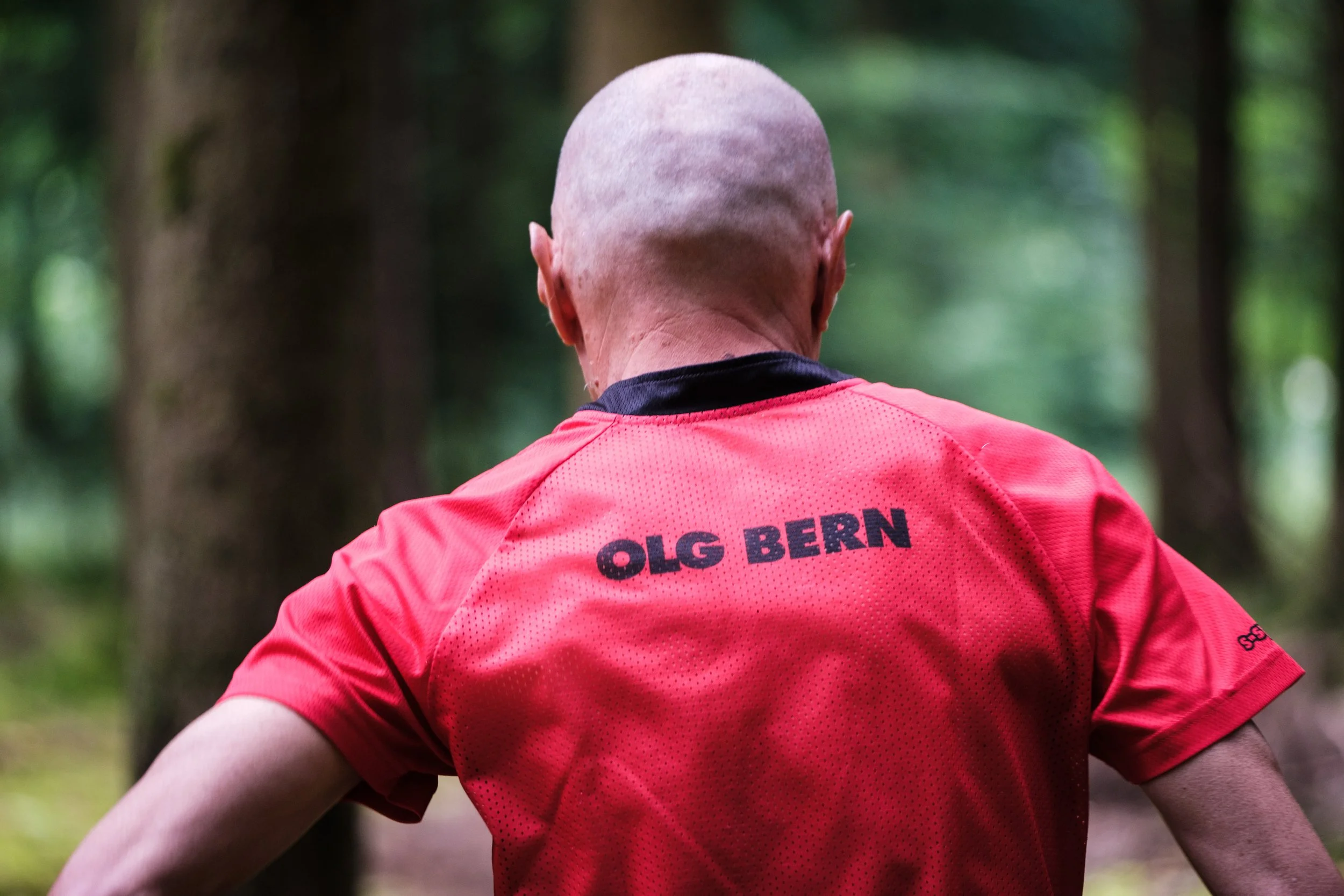 Rückansicht eines Mannes in einem roten Laufshirt mit der Aufschrift 'OLG BERN' im Wald.