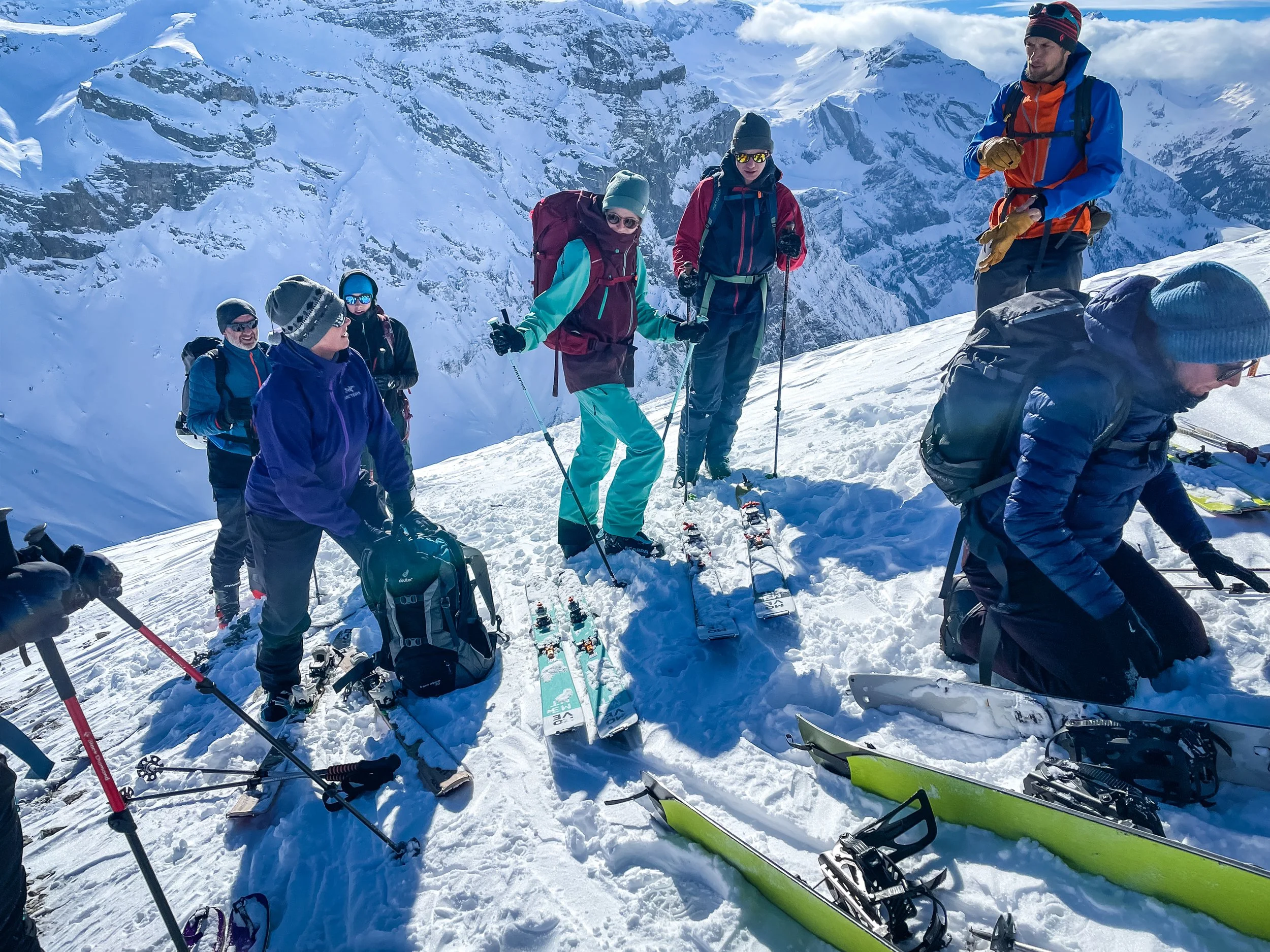 Eine Gruppe von Skitouristen auf einer schneebedeckten Bergkuppe, umgeben von hohen, schneebedeckten Bergen unter blauem Himmel. Sie sind mit Skibekleidung, Rucksäcken und Skistöcken ausgestattet.