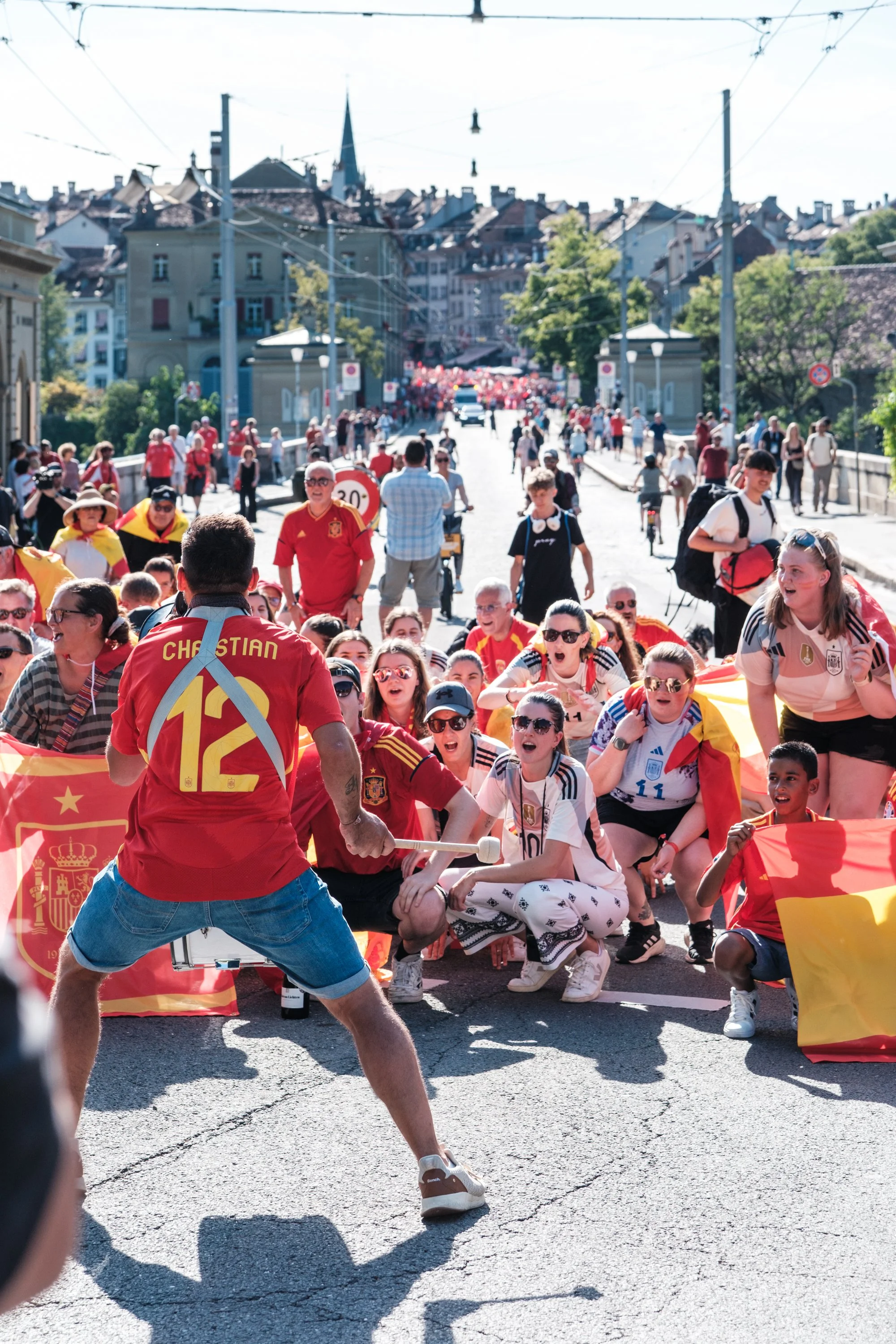 Gruppe von Menschen, die die spanische Fußballnationalmannschaft feiern, mit Flaggen und Trikots, auf einer Straße in einer Stadt bei sonnigem Wetter.