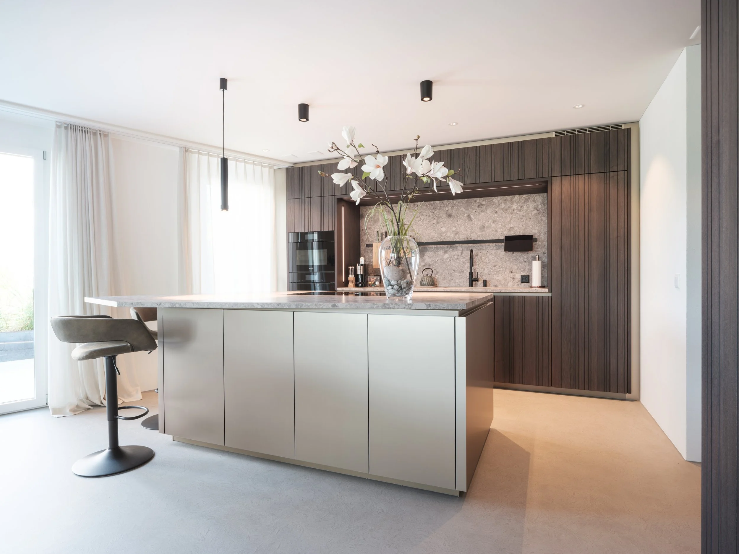 Moderne Küche mit Kücheninsel, dunklem Holzofen, grauer Wandverkleidung, und weißen Vorhängen bei Tageslicht.