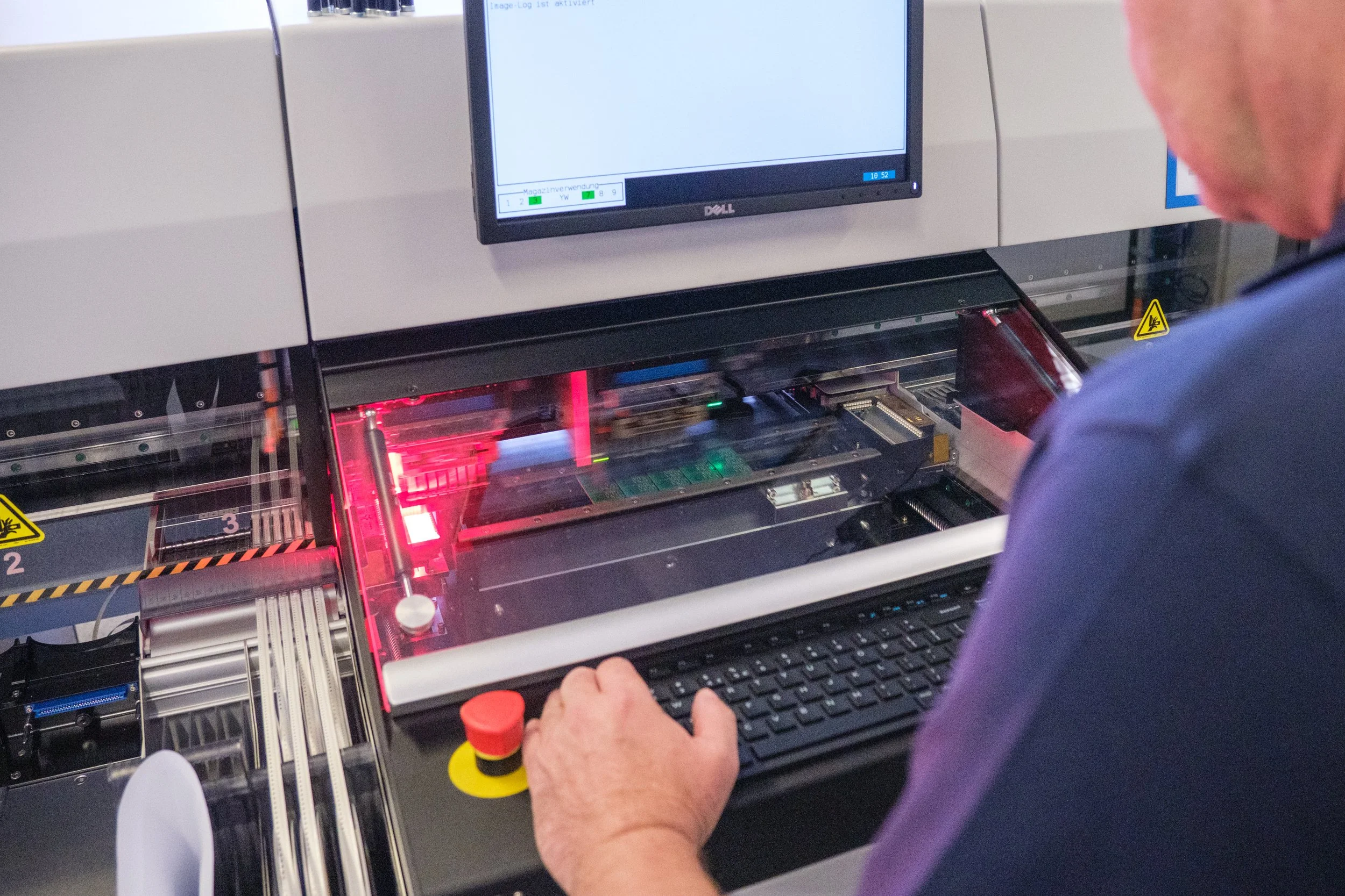Mensch arbeitet an einer elektronischen Maschine mit Computerbildschirm und rotem Laserlicht, vermutlich in einer Fabrik oder einem Labor.