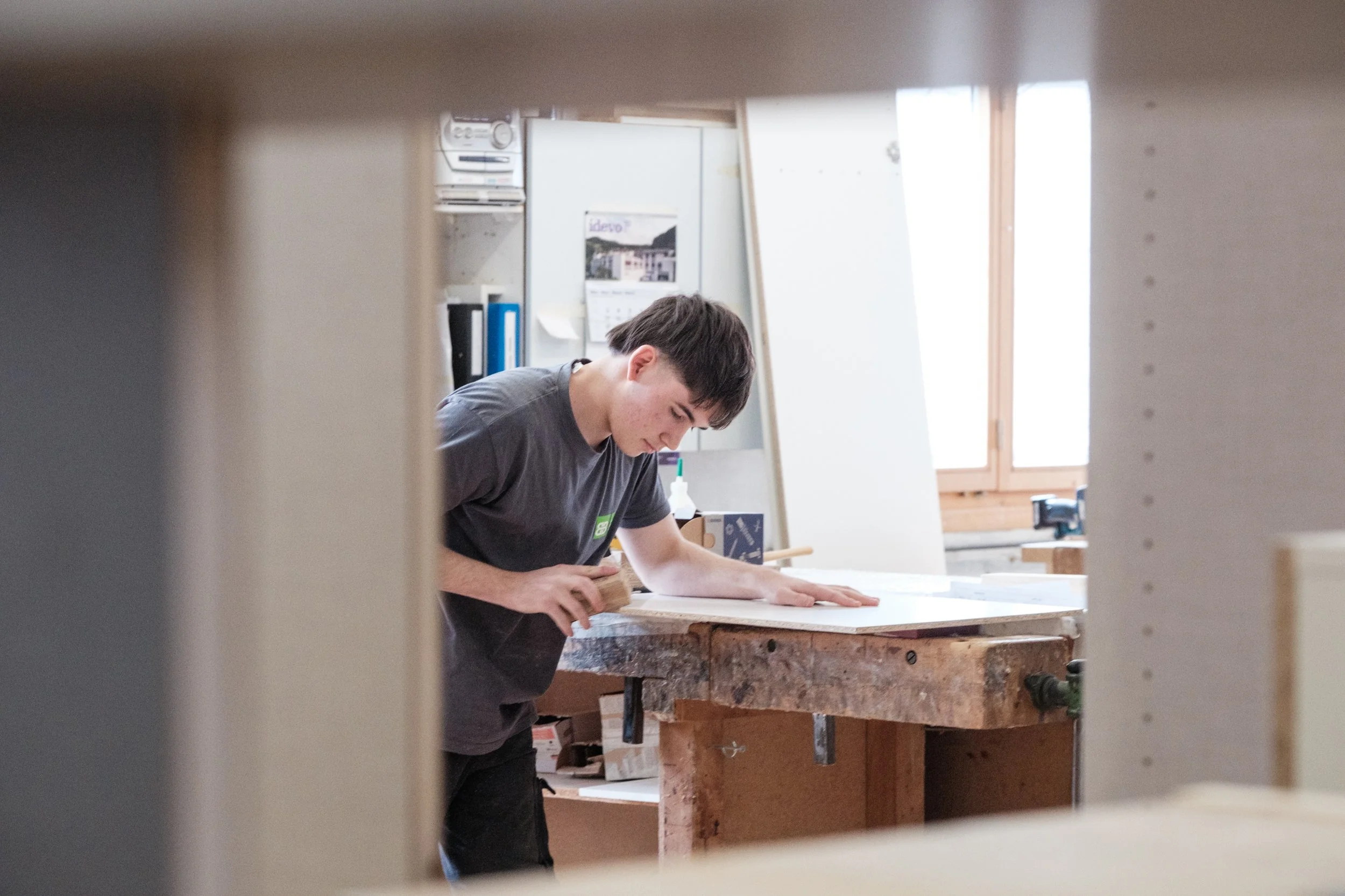 Junge arbeitet in einer Werkstatt an einer Holzarbeit, gesehen durch eine kleine Öffnung.