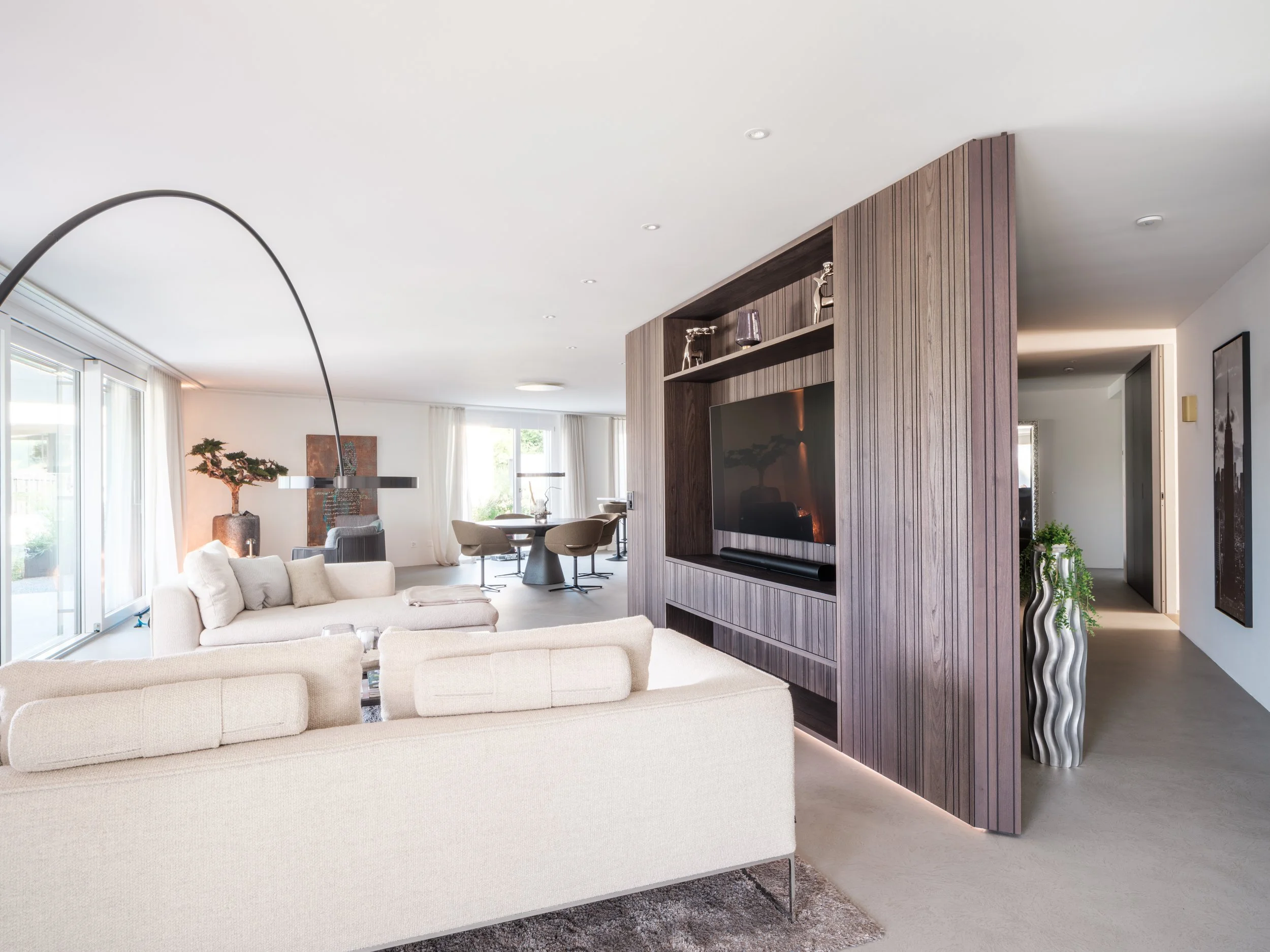 Moderner, heller Wohnbereich mit Sofa, Fernseher, Holzwand, Couchtisch, Essbereich mit runden Tischen und Stühlen, dekorative Pflanzen, große Fenster mit weißen Vorhängen.