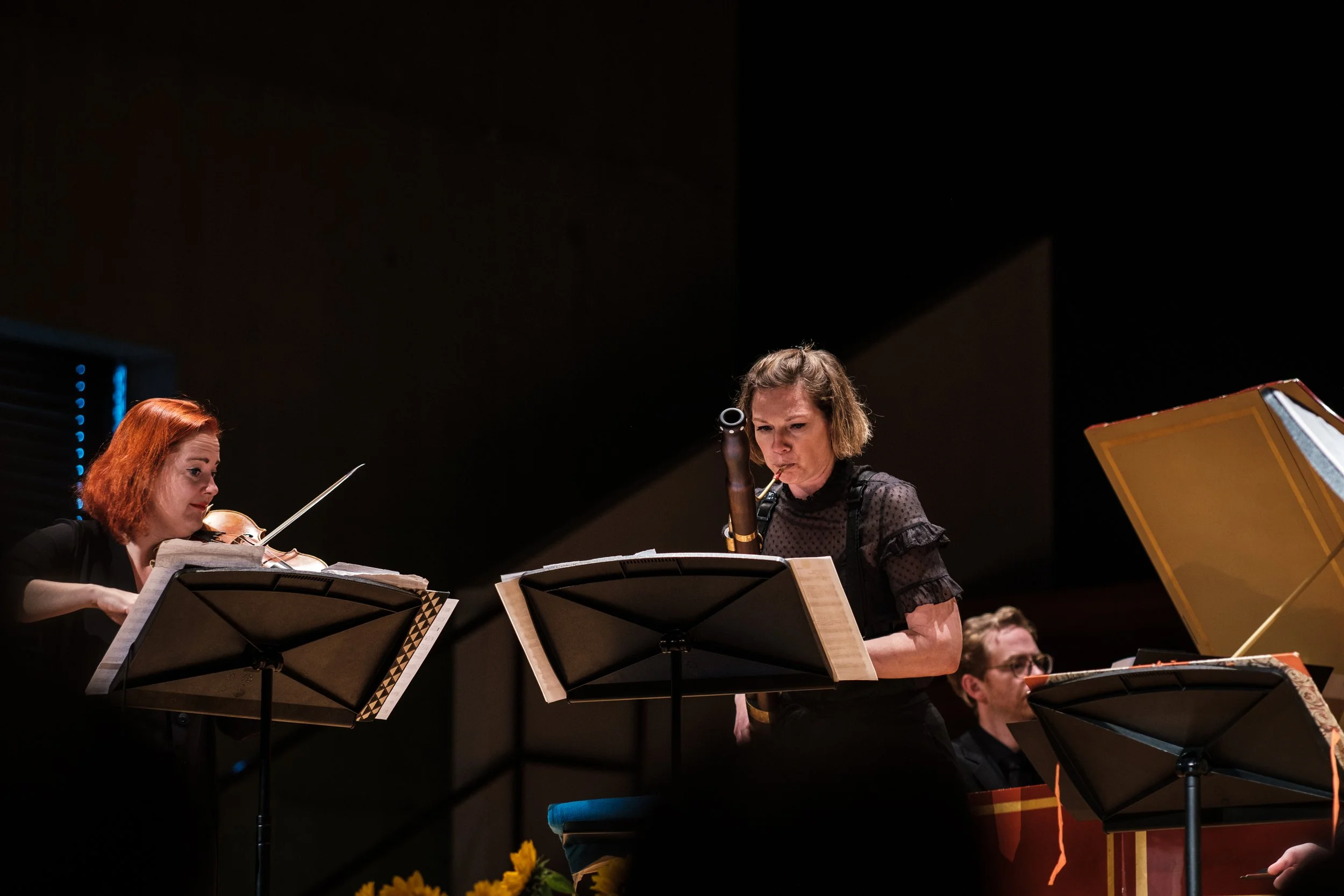 Musiker in einem Orchester spielen auf einer Bühne, eine Frau spielt die Violine, eine Frau spielt die Bassklarinette, ein Mann im Hintergrund spielt das Klavier.