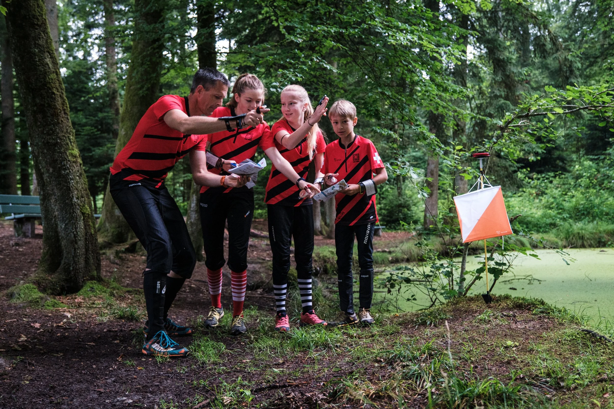 Vier Kinder und ein Erwachsener bei einer Geocaching-Aktivität im Wald, sie betrachten einen GPS-Tracker und eine Karte, alle tragen gleiche rote und schwarz gestreifte T-Shirts.