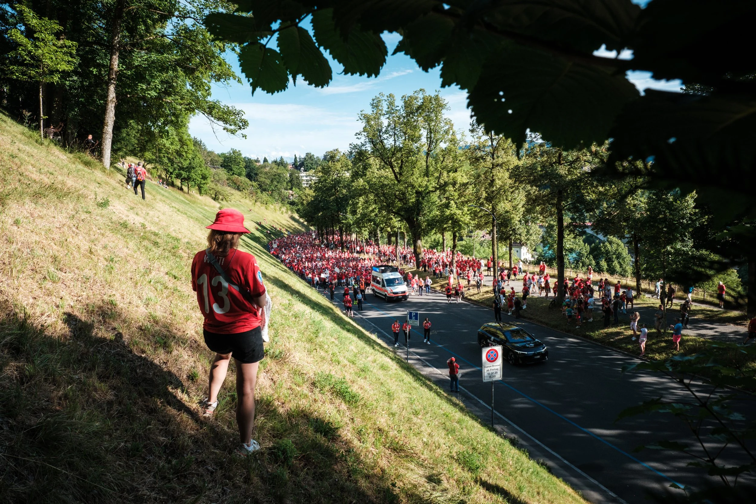 Ein Mädchen in rotem Trikot und rotem Hut steht an einem Hang und schaut auf eine Menschenmenge, die sich auf einer Straße versammelt hat, vermutlich für ein Fest oder eine Parade, umgeben von Bäumen bei schönem Wetter.