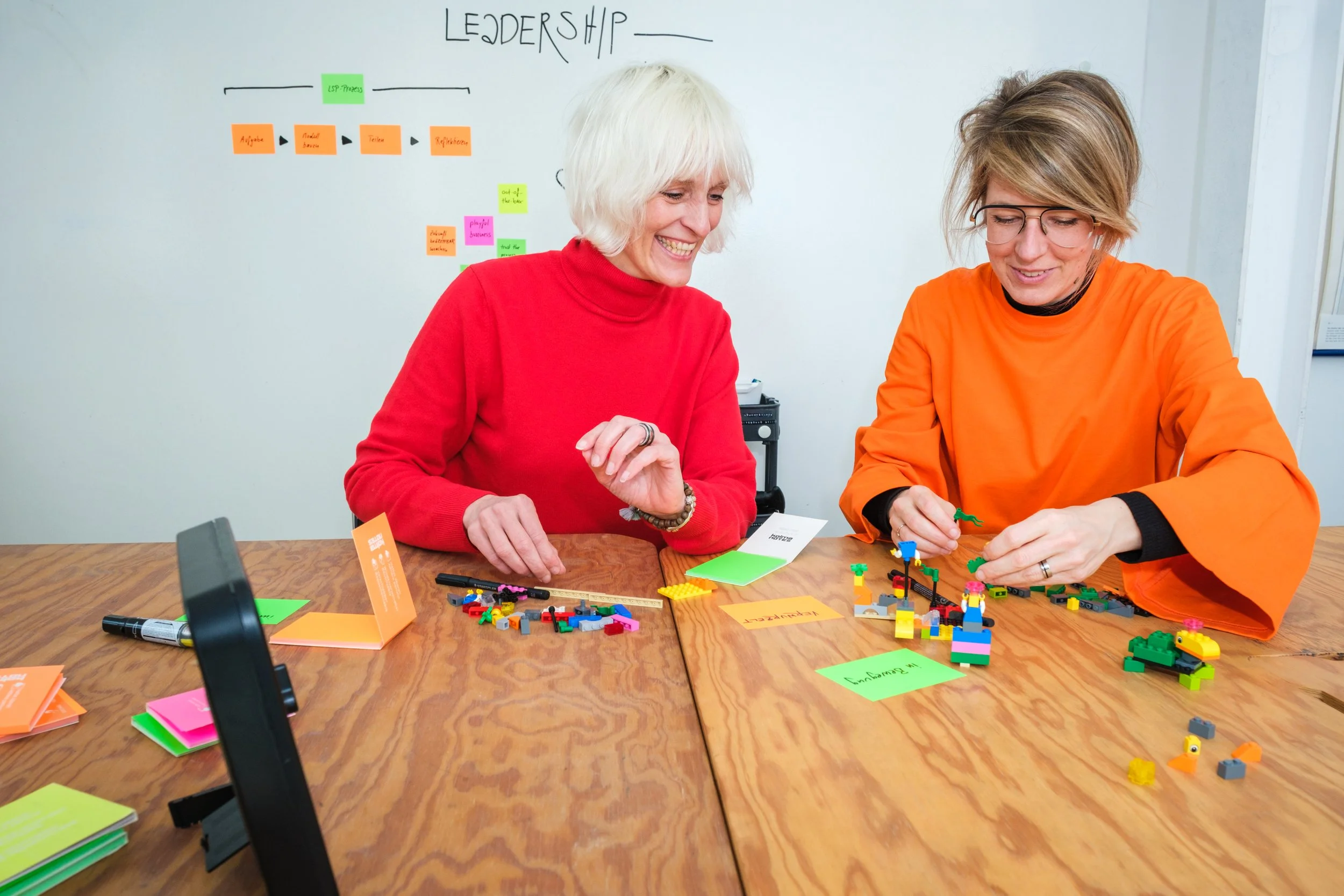 Zwei Frauen spielen gemeinsam mit LEGO-Steinen auf einem Holztisch. Im Hintergrund steht eine Whiteboard mit farbigen Post-its und einem Diagramm zum Thema Leadership.