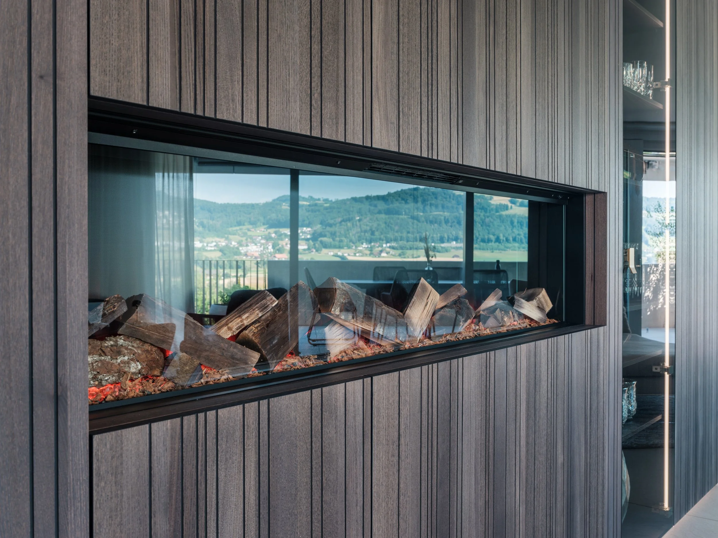 Moderne, eingebaute Feuerstelle mit Holzscheiten, umgeben von aufwändig gearbeiteter dunkler Holzverkleidung, mit Blick auf eine grüne Hügellandschaft durch große Fenster
