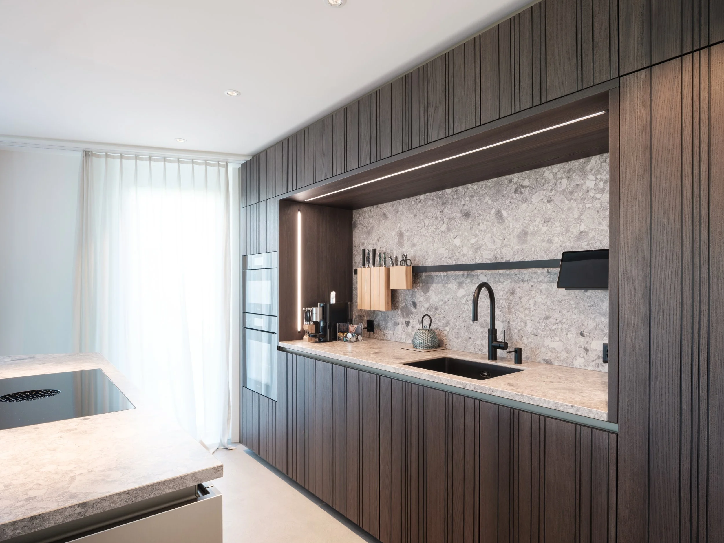 Moderne Küchenschränke mit dunklem Holz, helle Arbeitsplatte, graue Marmorrückwand, eingebautem Wasserhahn und Küchenutensilien, viel natürlichem Licht durch große Fenster mit weißen Vorhängen.