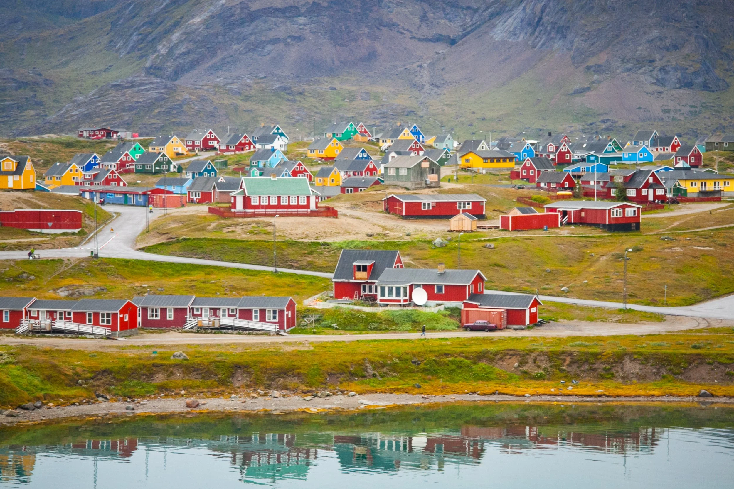 Bunte Häuser an einem See in einer bergigen Landschaft, größtenteils in Rot, blau, gelb, grün und grau, mit Wiesen und Wegen