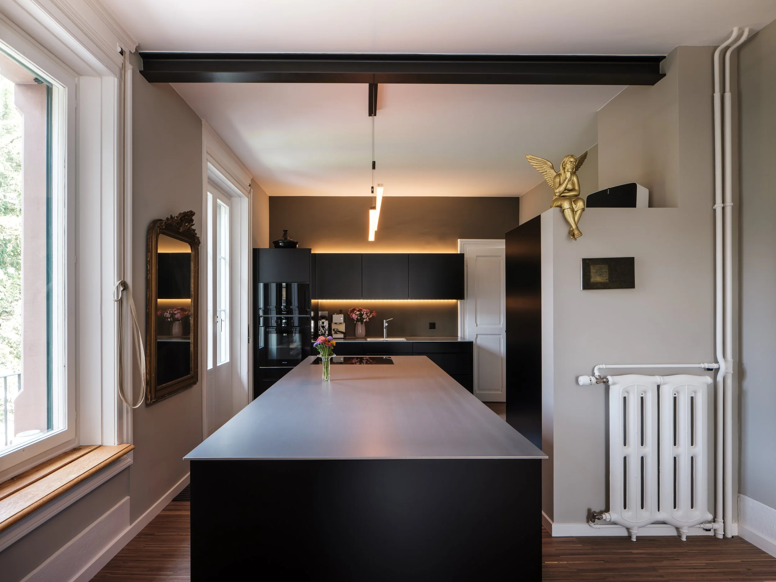 Moderne Küche mit schwarzen Schränken, langer Kücheninsel mit Blumenstrauß, weißen Wänden, großen Fenstern und einem antiken Spiegel an der Wand.