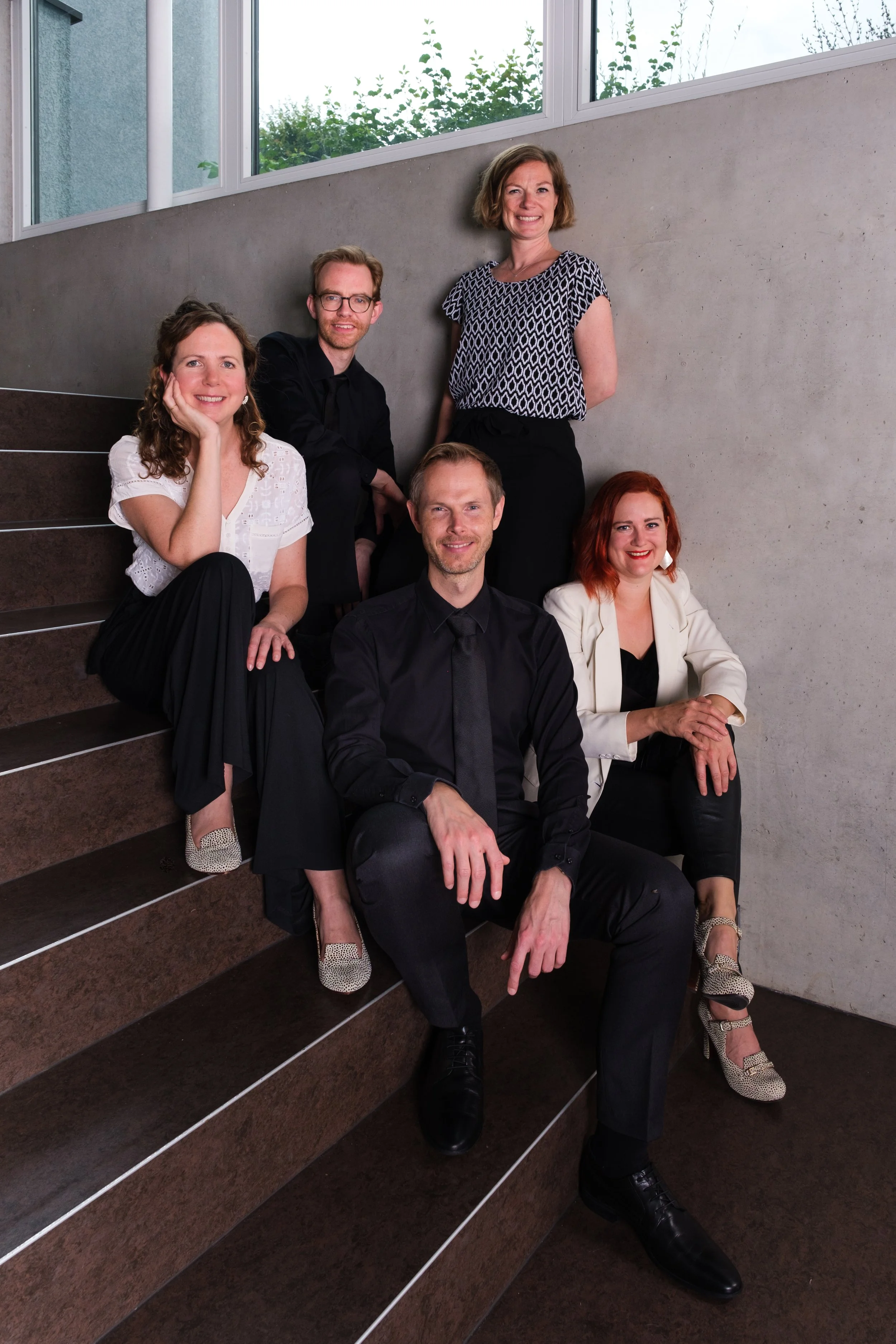 Sechs Menschen, drei Frauen und drei Männer, sitzen und stehen auf einer Treppe in einem modernen Gebäude, im Hintergrund Fenster und Bäume sichtbar.