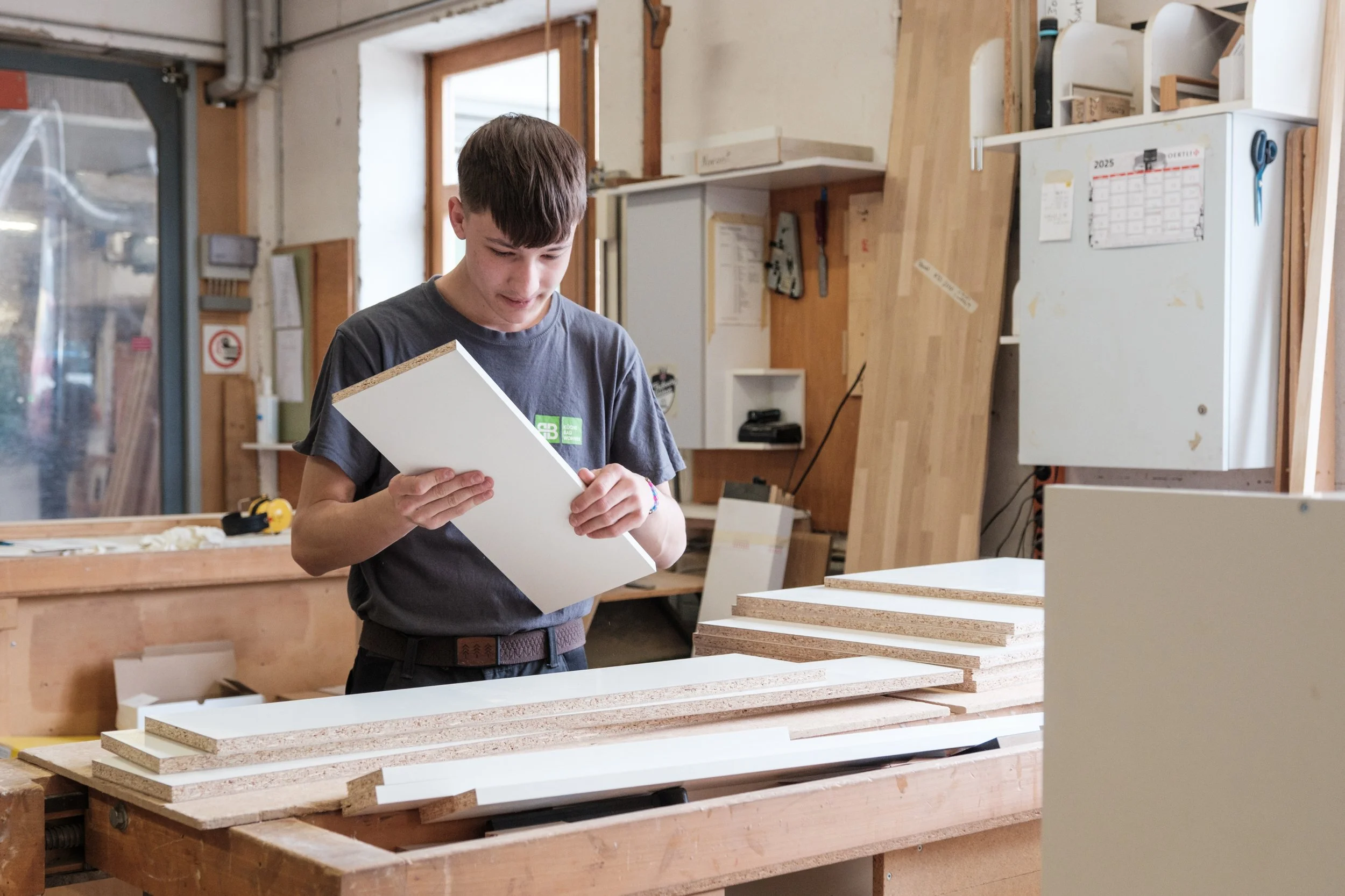 Ein junger Mann in einer Werkstatt, der eine weiße Holzplatte betrachtet, mit mehreren weiteren Holzplatten auf einem Arbeitstisch. Die Werkstatt hat Holzregale und Werkzeuge an den Wänden.
