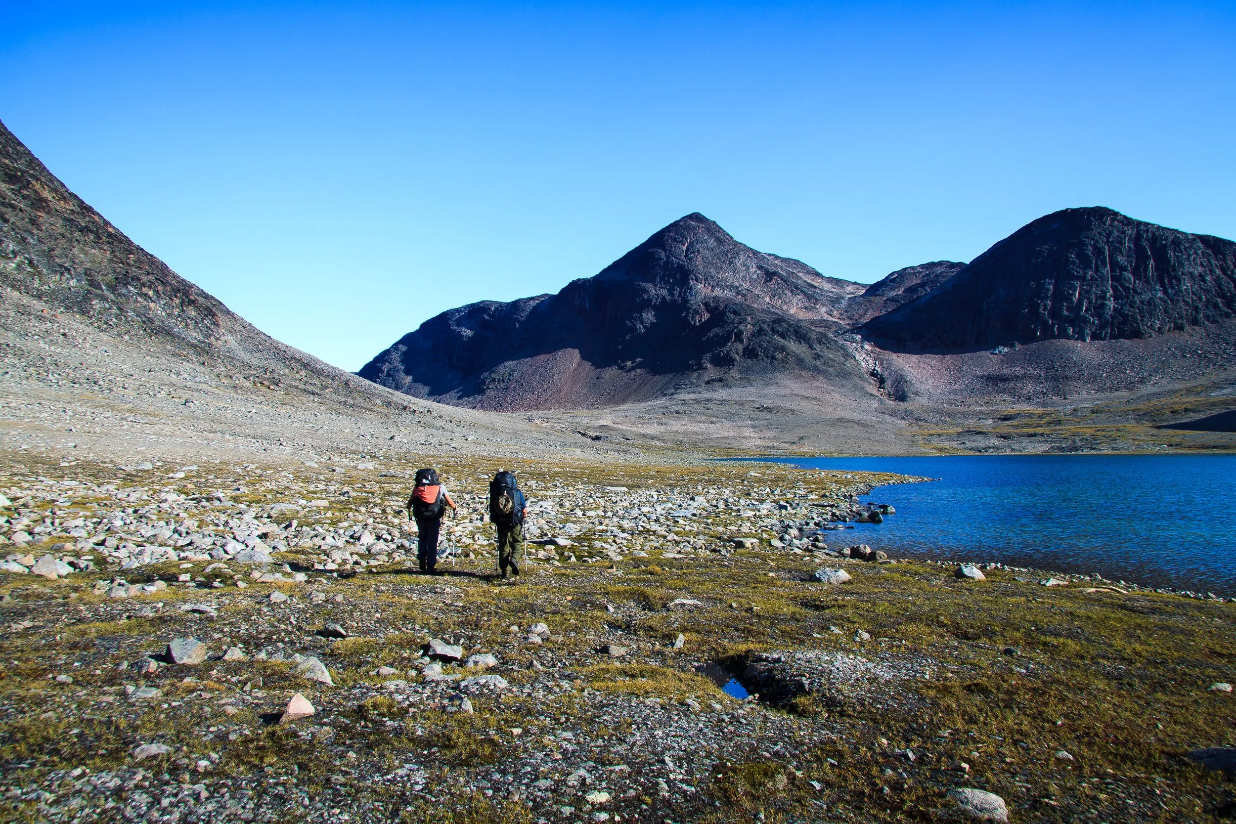 Zwei Wanderer mit Rucksäcken unterwegs in einer Berglandschaft mit See, steilen Bergen und klarem Himmel