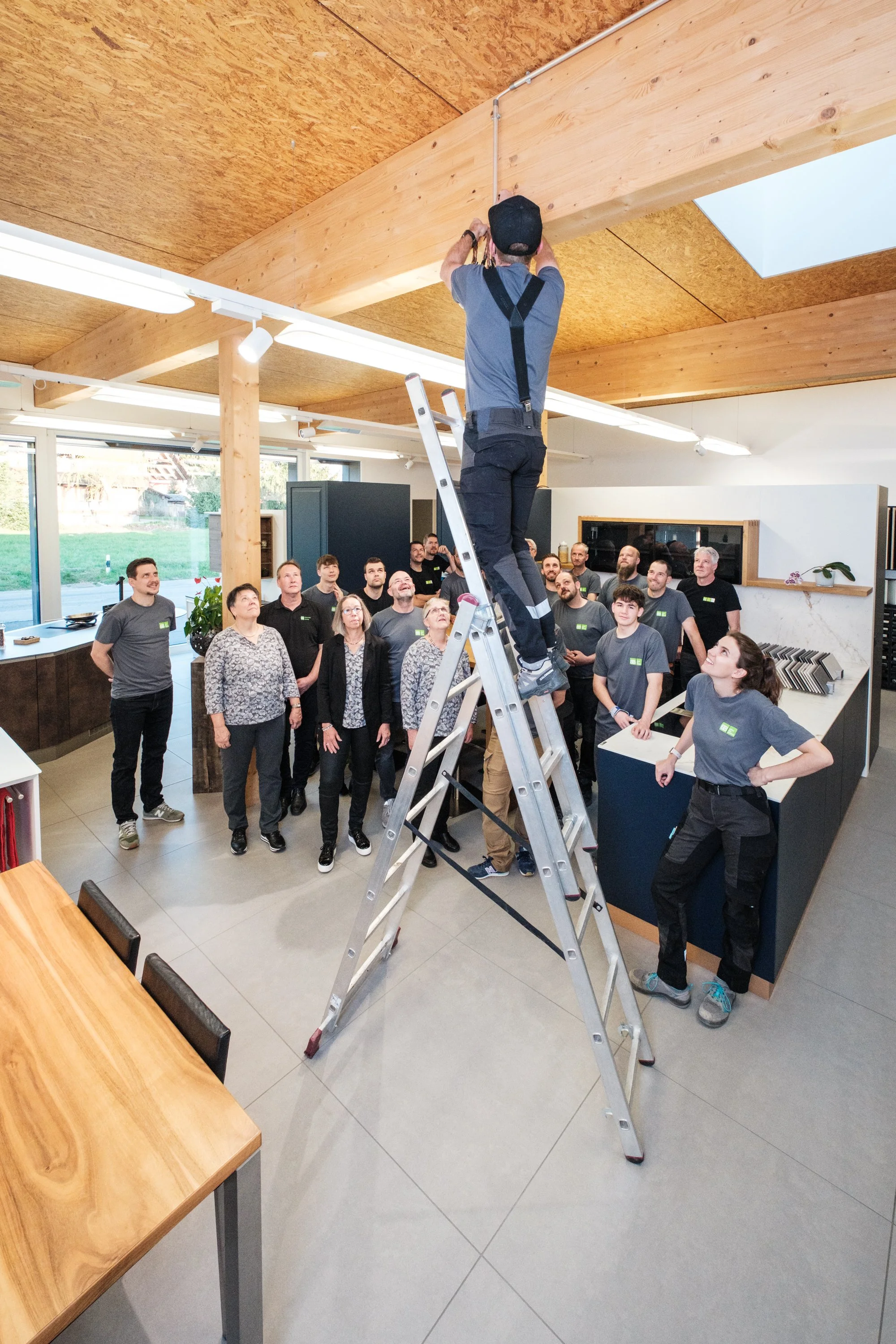 Eine Gruppe von Menschen beobachtet, wie ein Techniker auf einer Leiter eine elektrische Leitung am Deckenbalken in einem modernen Raum befestigt, während alle zuschauen.