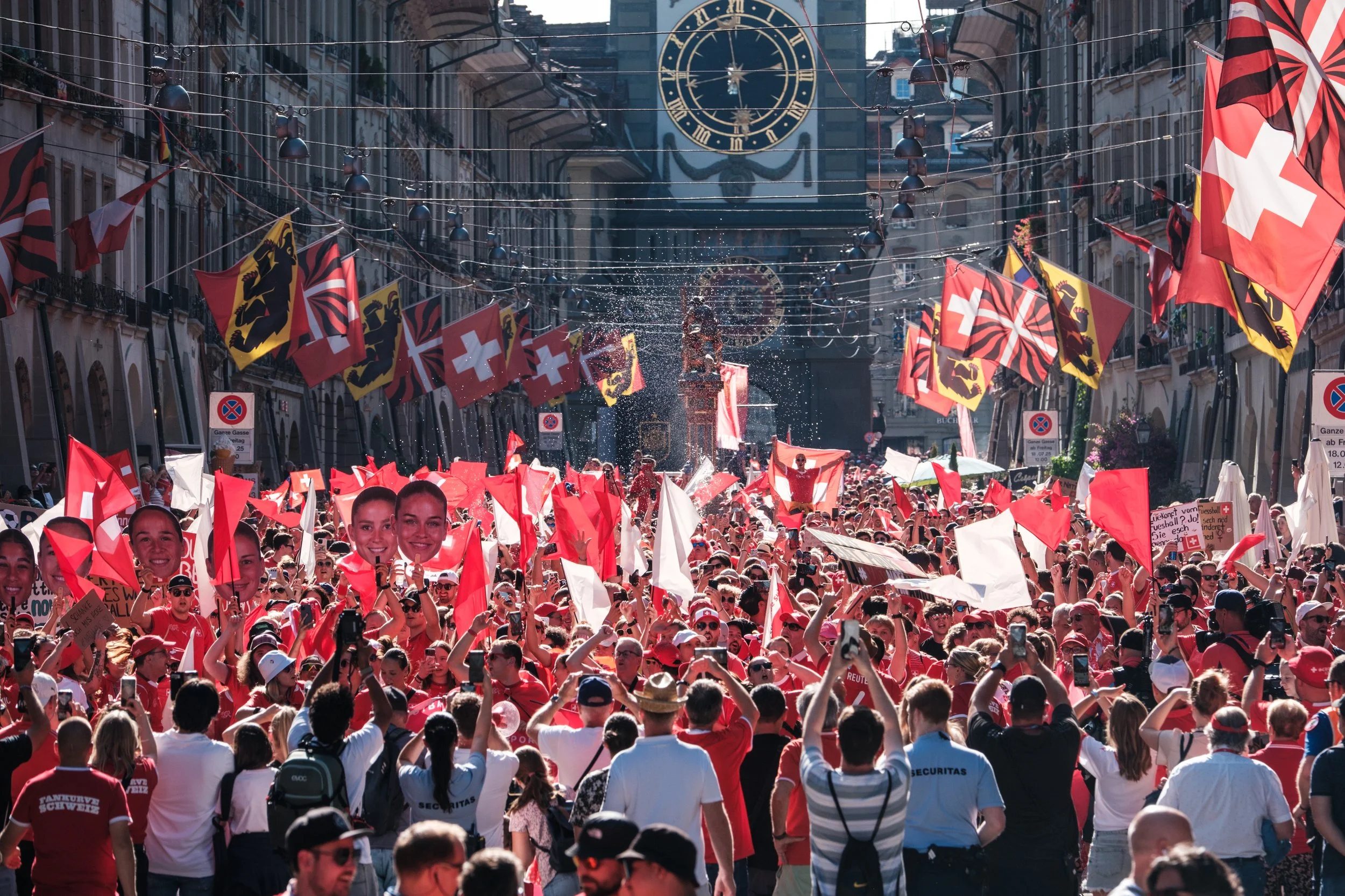 Menschenmenge bei einer Demonstration in der Stadt, viele Menschen halten Schweizerflaggen und Plakate mit Fotos, darunter einige mit lachenden Gesichtern, im Hintergrund ist eine große Uhr und das Stadtbild zu sehen.