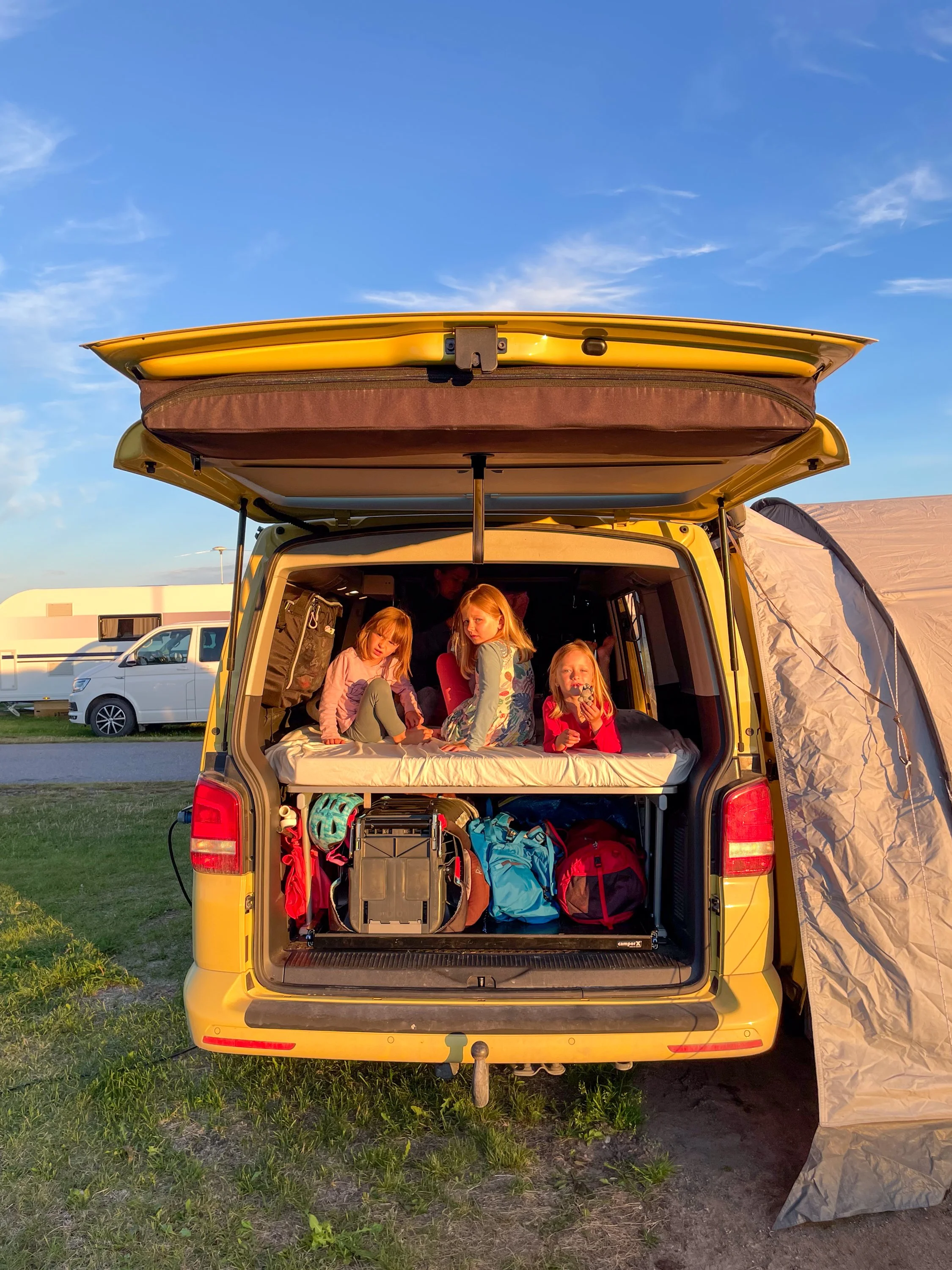 Drei Kinder sitzen auf einer Matratze im geöffneten Kofferraum eines gelben Wohnmobils, umgeben von Rucksäcken, bei Sonnenuntergang auf einem Campingplatz.
