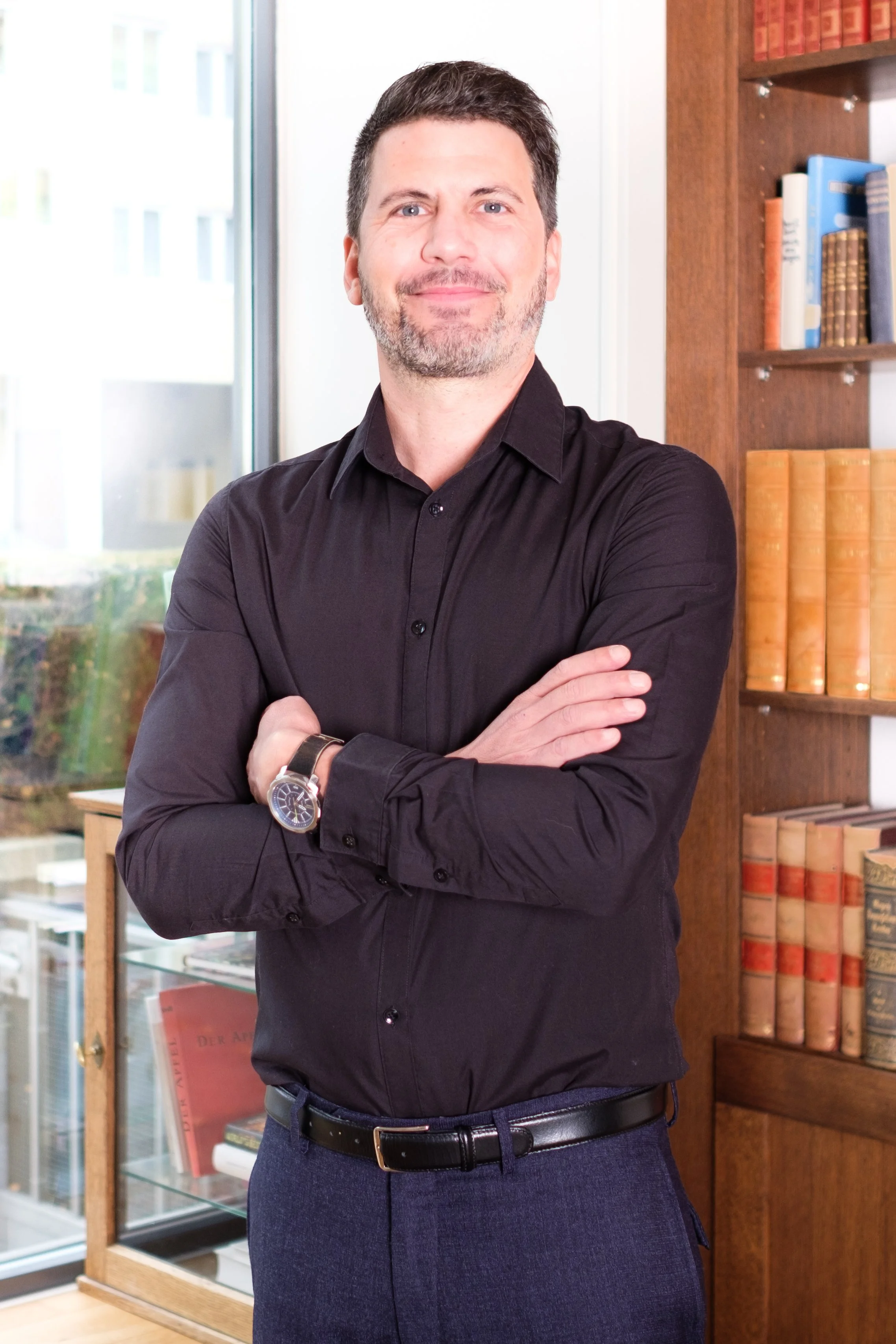 Ein Mann mit dunklem Hemd und Armbanduhr steht mit verschränkten Armen in einem Raum mit Bücherregalen und Fenster.