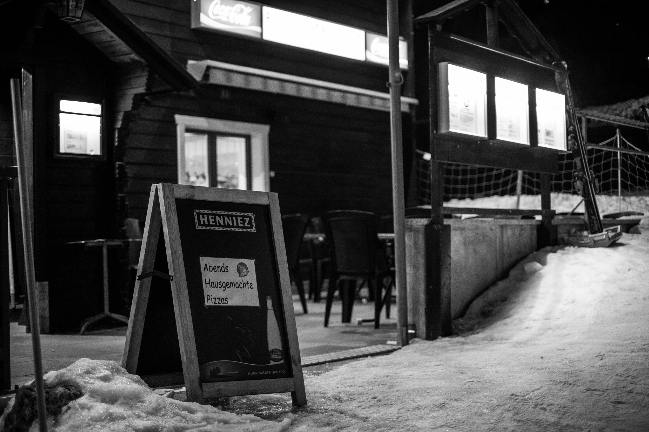 Ein schwarzes Pult im Schnee vor einem Restaurant mit Menüanzeigen. Das Pult bewirbt abends hausgemachte Pizzas. Es ist Nacht, und die Umgebung ist verschneit.