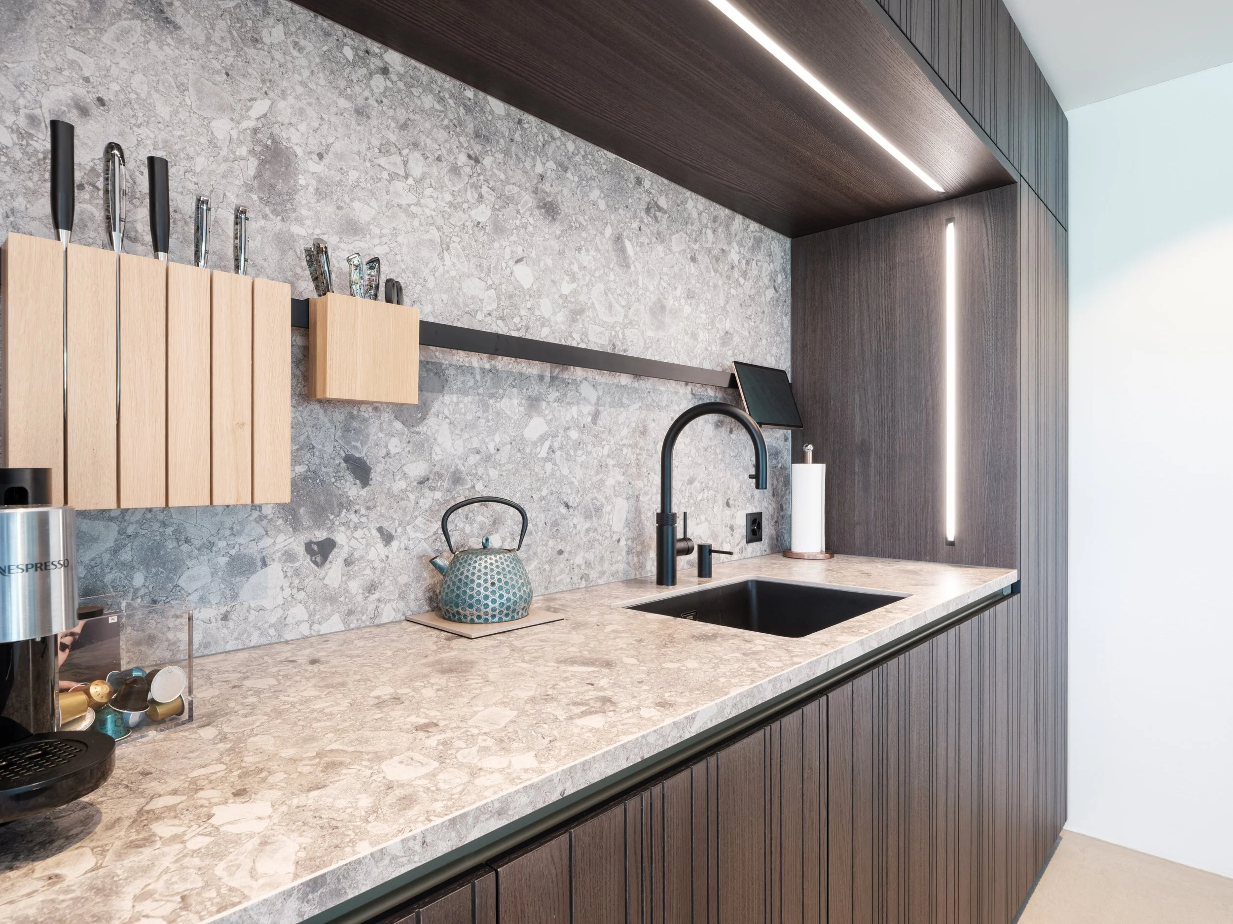 Moderne Küchenzeile mit schwarzen Armaturen, Marmorbotschaft und dunklen Holzschränken, Küchenutensilien an der Wand, Teekanne auf der Arbeitsplatte, Kaffeeautomat.