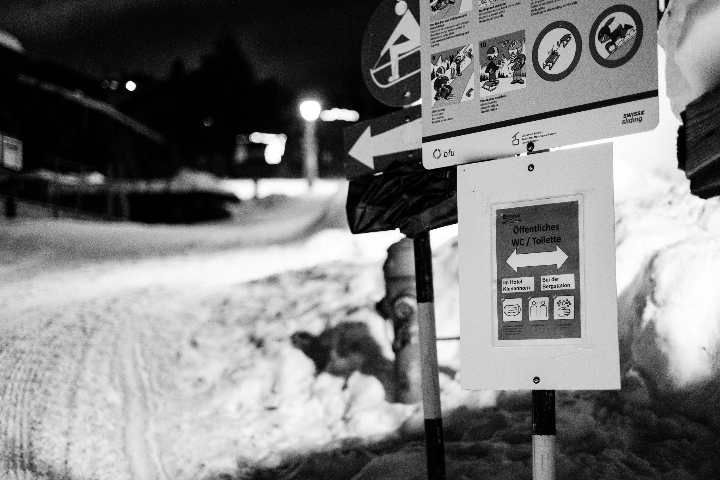 Hinweisschilder am Schneepflug, die den Weg zu öffentlichen Toiletten im Hotel Klenenhorn und bei der Bergstation in einem Schneelandschaft bei Nacht anzeigen.