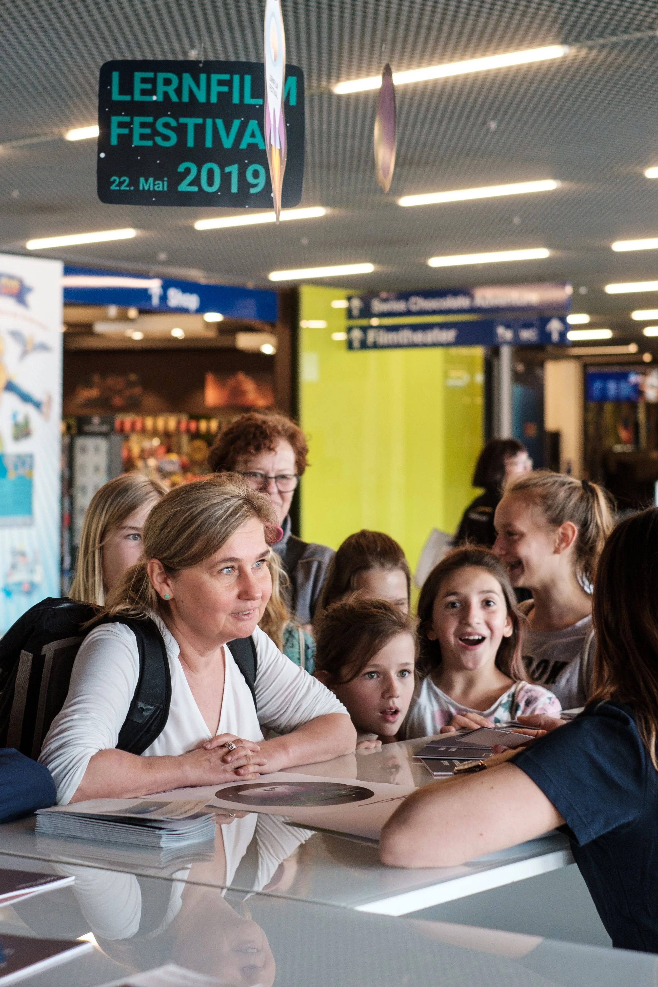 Eine Gruppe von Menschen, darunter mehrere Kinder und Erwachsene, steht an einem Informationsschalter in einer öffentlichen Einrichtung, vermutlich einem Bahnhof oder Flughafen, mit einem digitalen Ankündigungsdisplay im Hintergrund, das den "Lernfil
