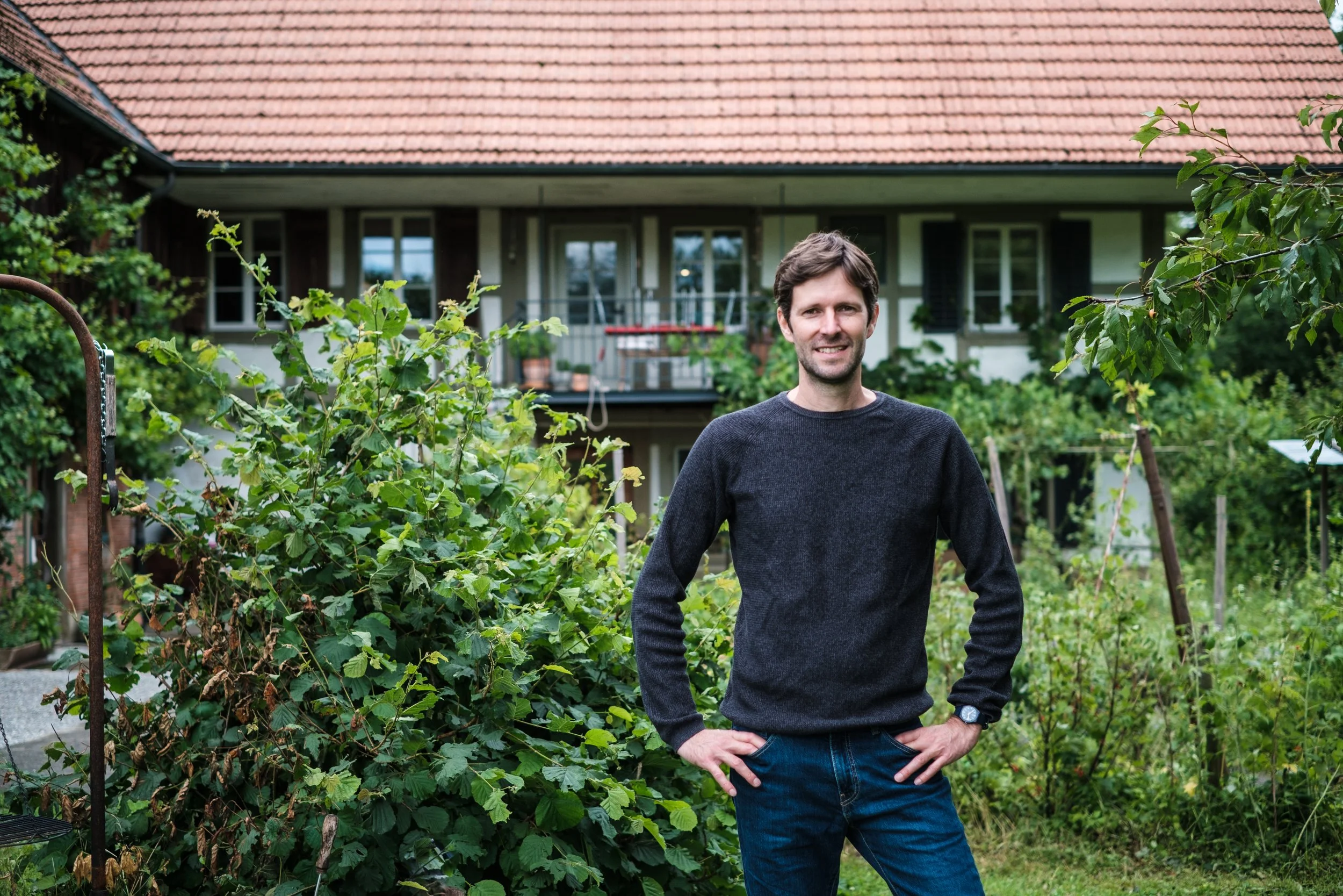 Ein Mann steht in einem Garten vor einem Haus mit Balkon und roten Dachziegeln, umgeben von grünen Pflanzen und Bäumen.