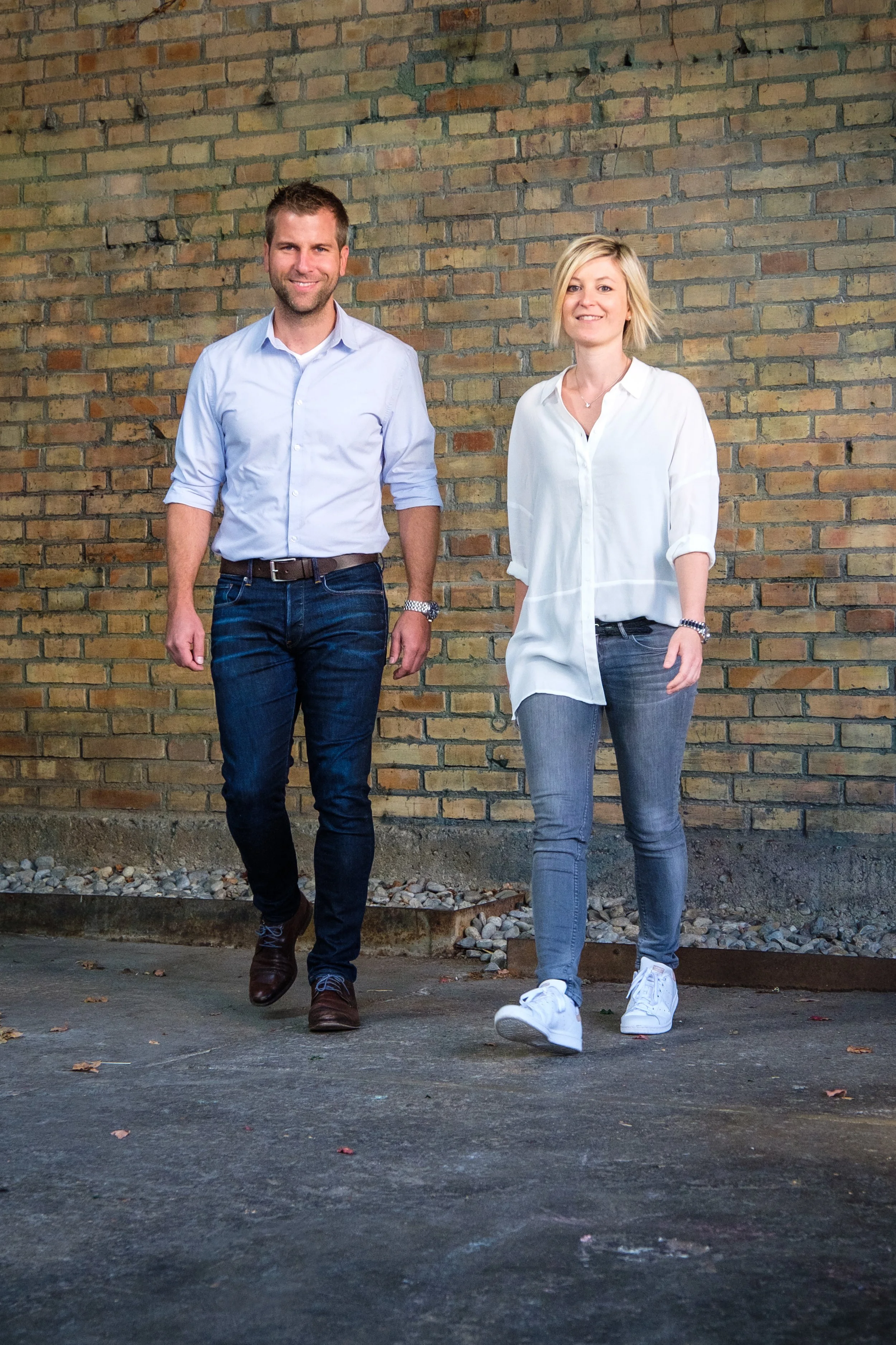 Zwei lachende Erwachsene, ein Mann und eine Frau, gehen nebeneinander vor einer Ziegelmauer. Der Mann trägt ein hellblaues Hemd und dunkle Jeans, die Frau ein weißes Hemd und graue Jeans. Sie scheinen entspannt und freundlich zu sein.