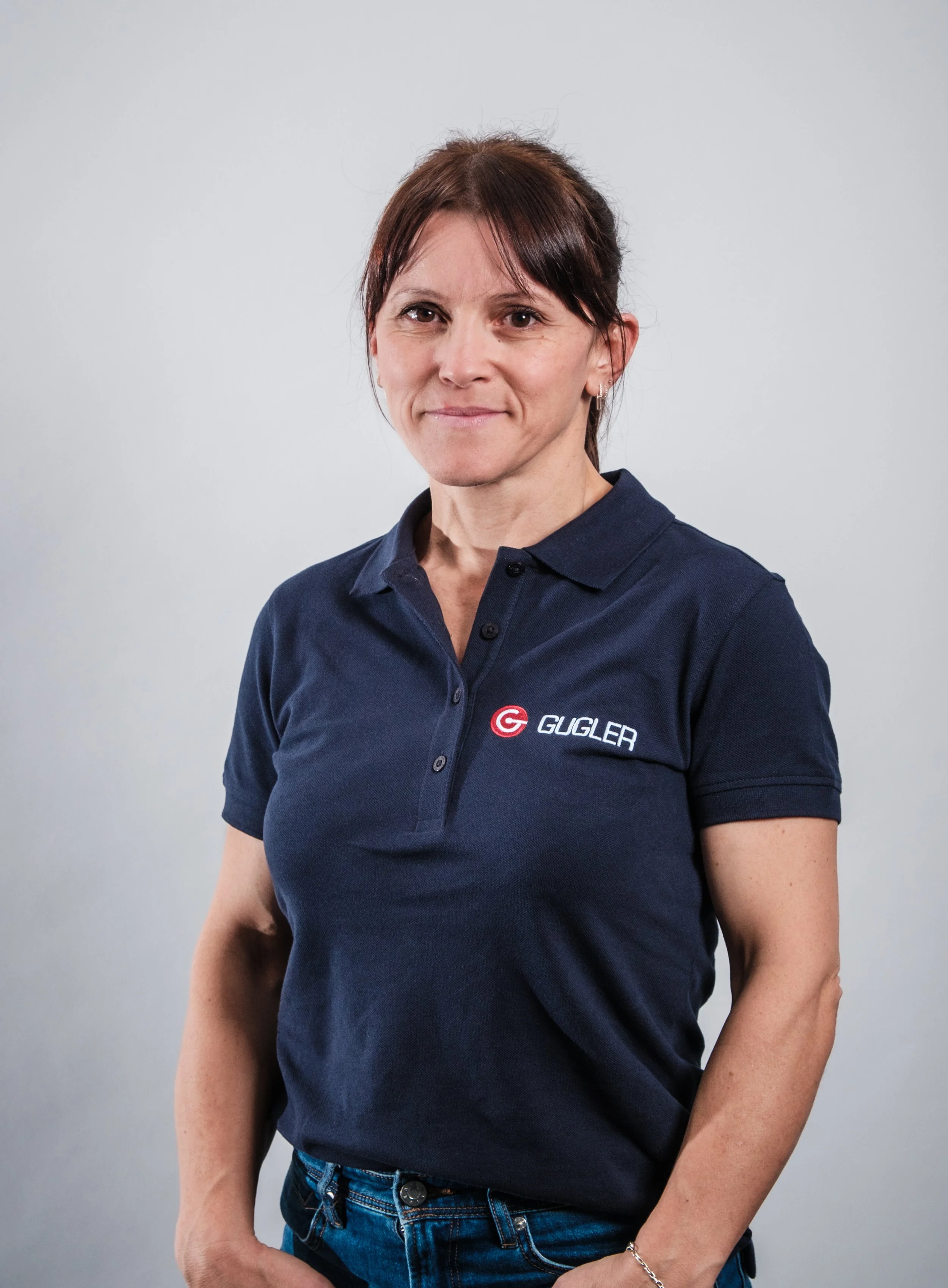 Eine Frau trägt ein dunkelblaues Polo-Shirt mit dem Logo und Namen 'Gugler'. Sie steht vor einem grauen Hintergrund und schaut in die Kamera.