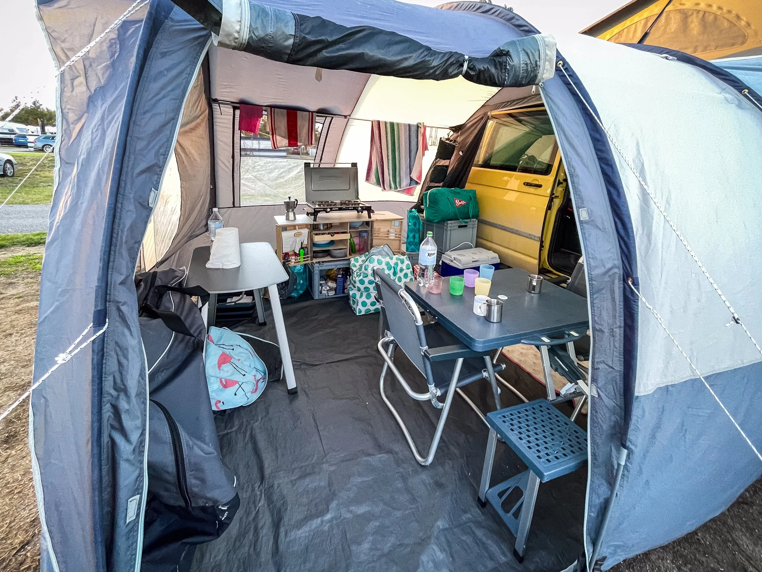 Innenansicht eines Zelt mit einem kleinen Kocher, Esstisch und Campingausrüstung, daneben ein gelber Van