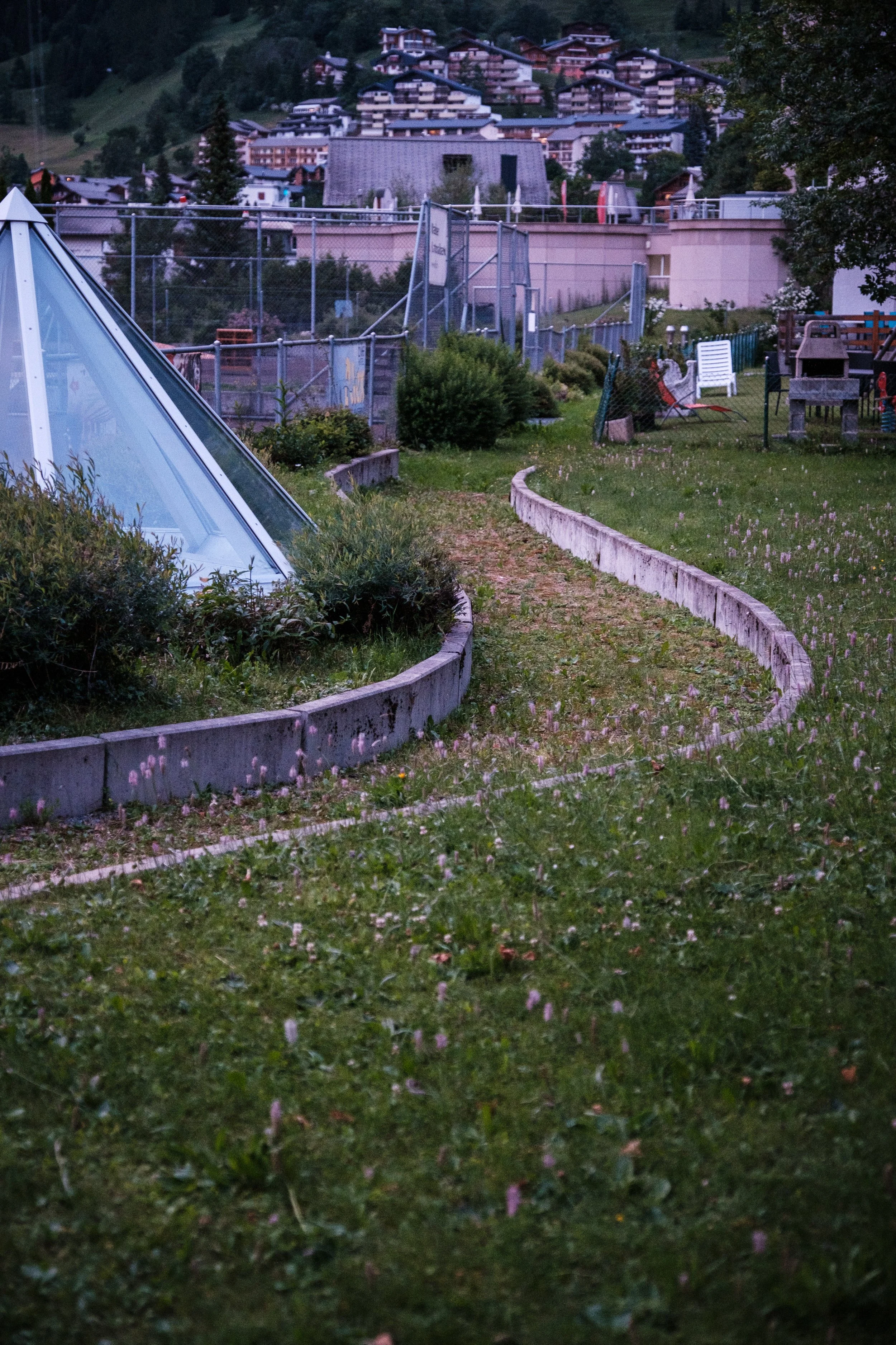 Blick auf einen Garten mit Weg, Glasgeometrie-Struktur und Gartenmöbeln, im Hintergrund Häuser auf einem Hügel.