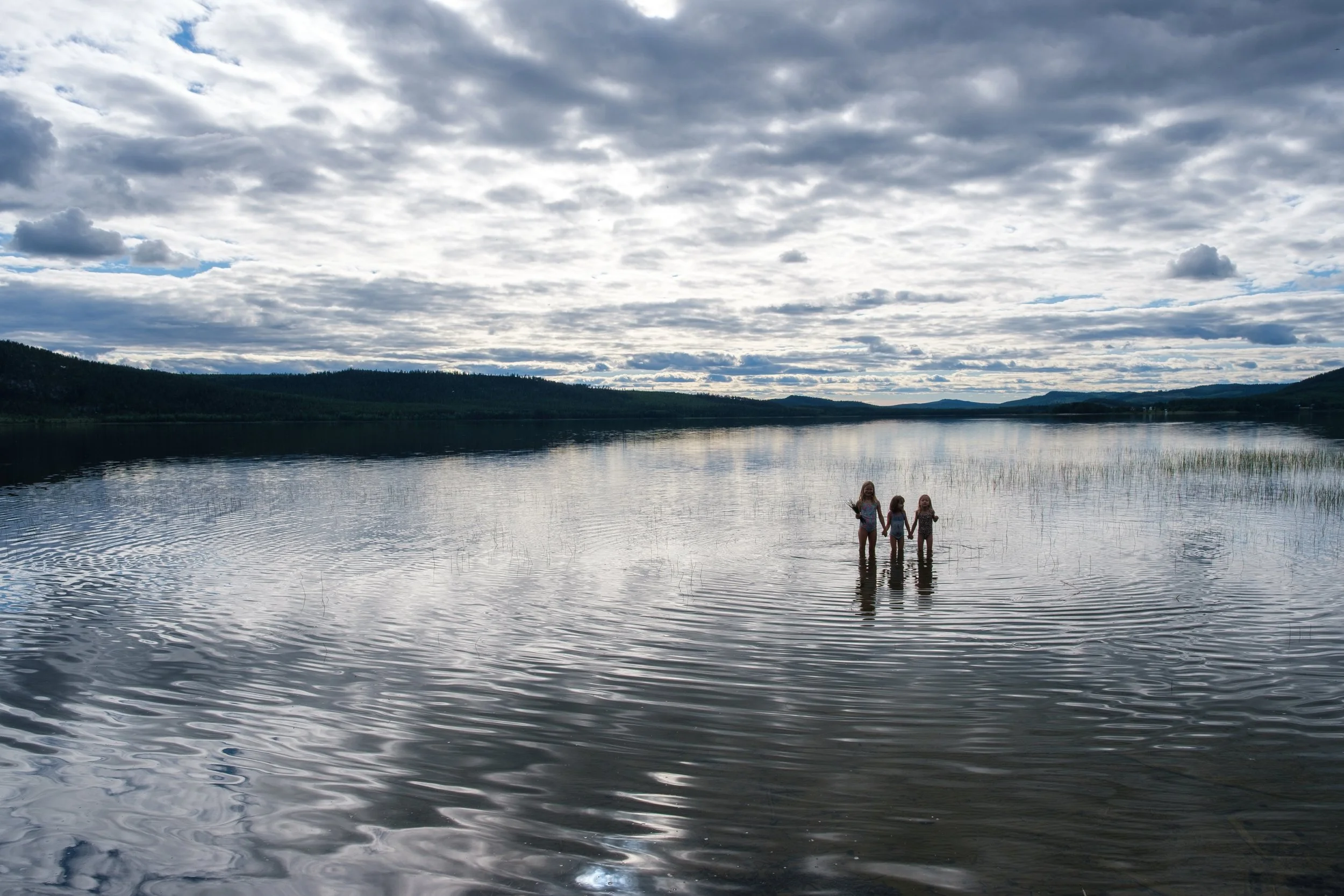 Drei Kinder stehen im Wasser eines Sees, halten sich an den Händen und betrachten die Landschaft unter einem bewölkten Himmel.